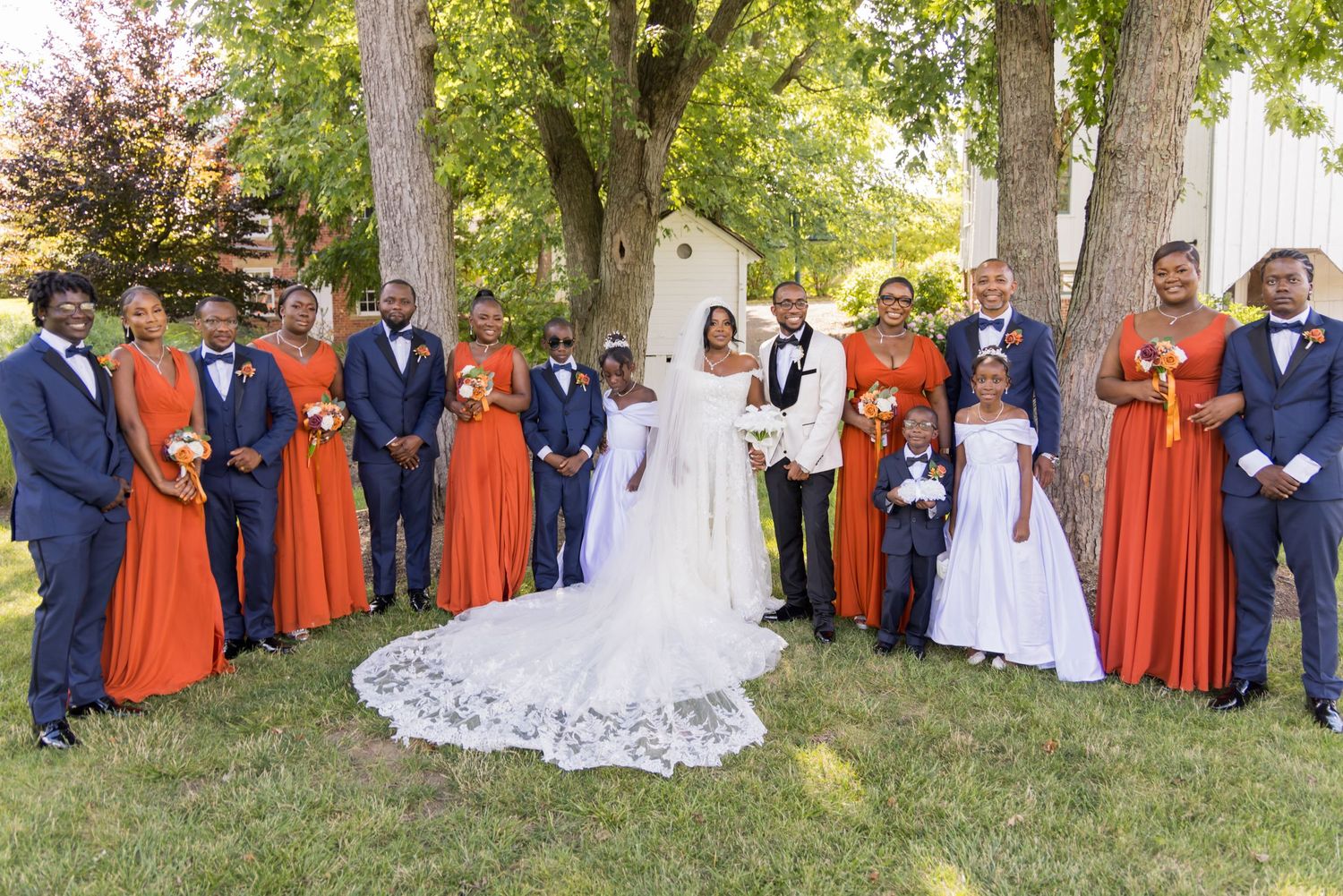 Wedding party in orange bridesmaid dresses and navy suits pose outdoors with bride in white gown.