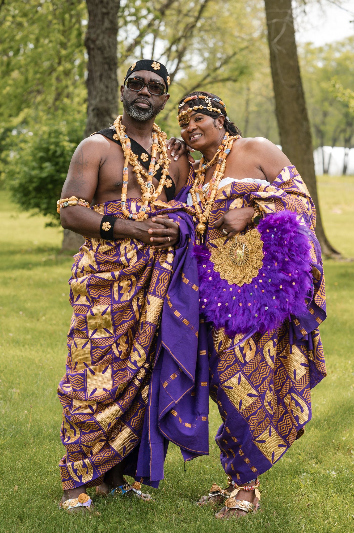 Couple wearing traditional African wedding attire poses together in a park setting.
