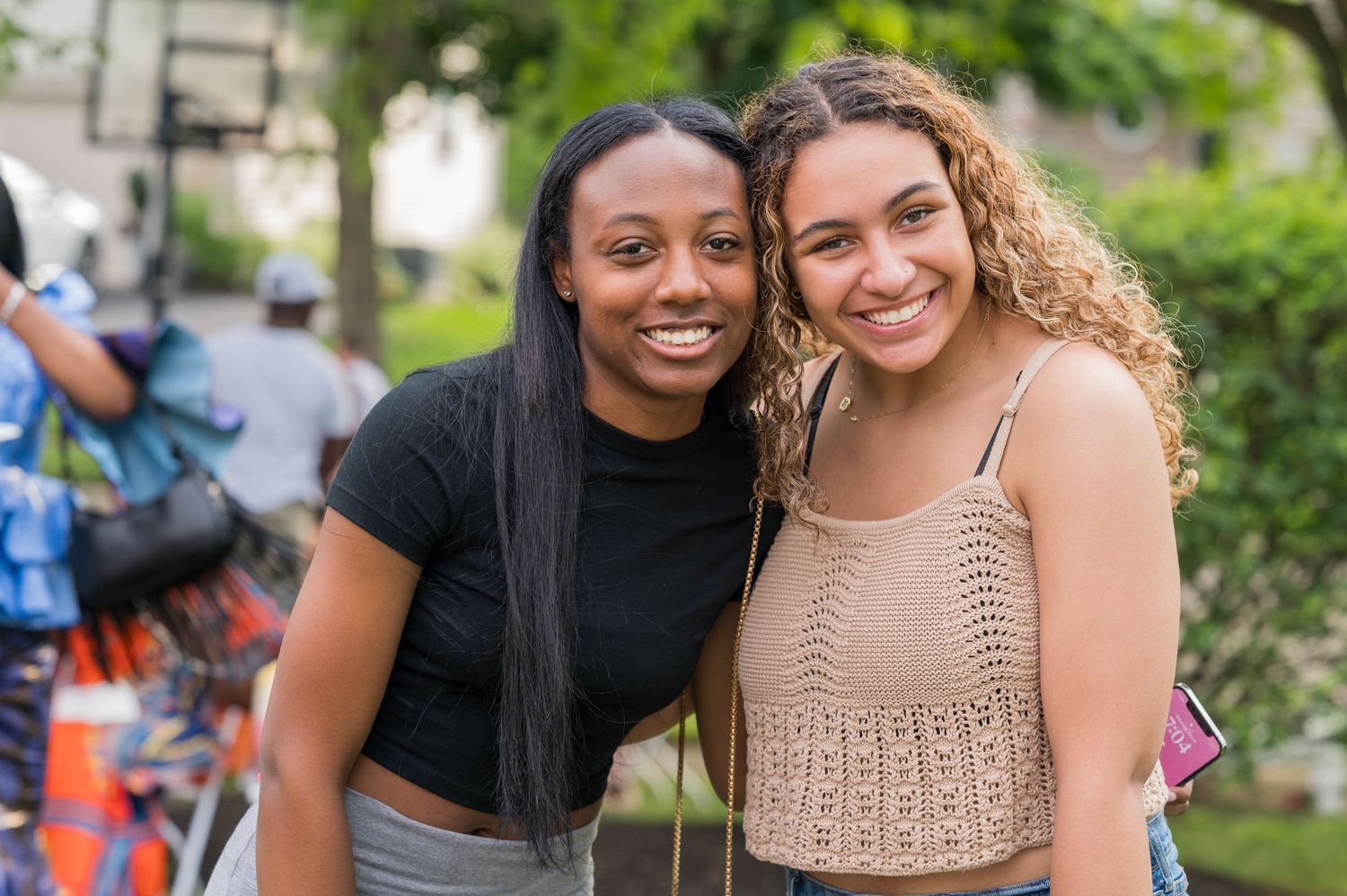 Two friends pose together outdoors on a sunny day with greenery and parked bikes visible in the background.