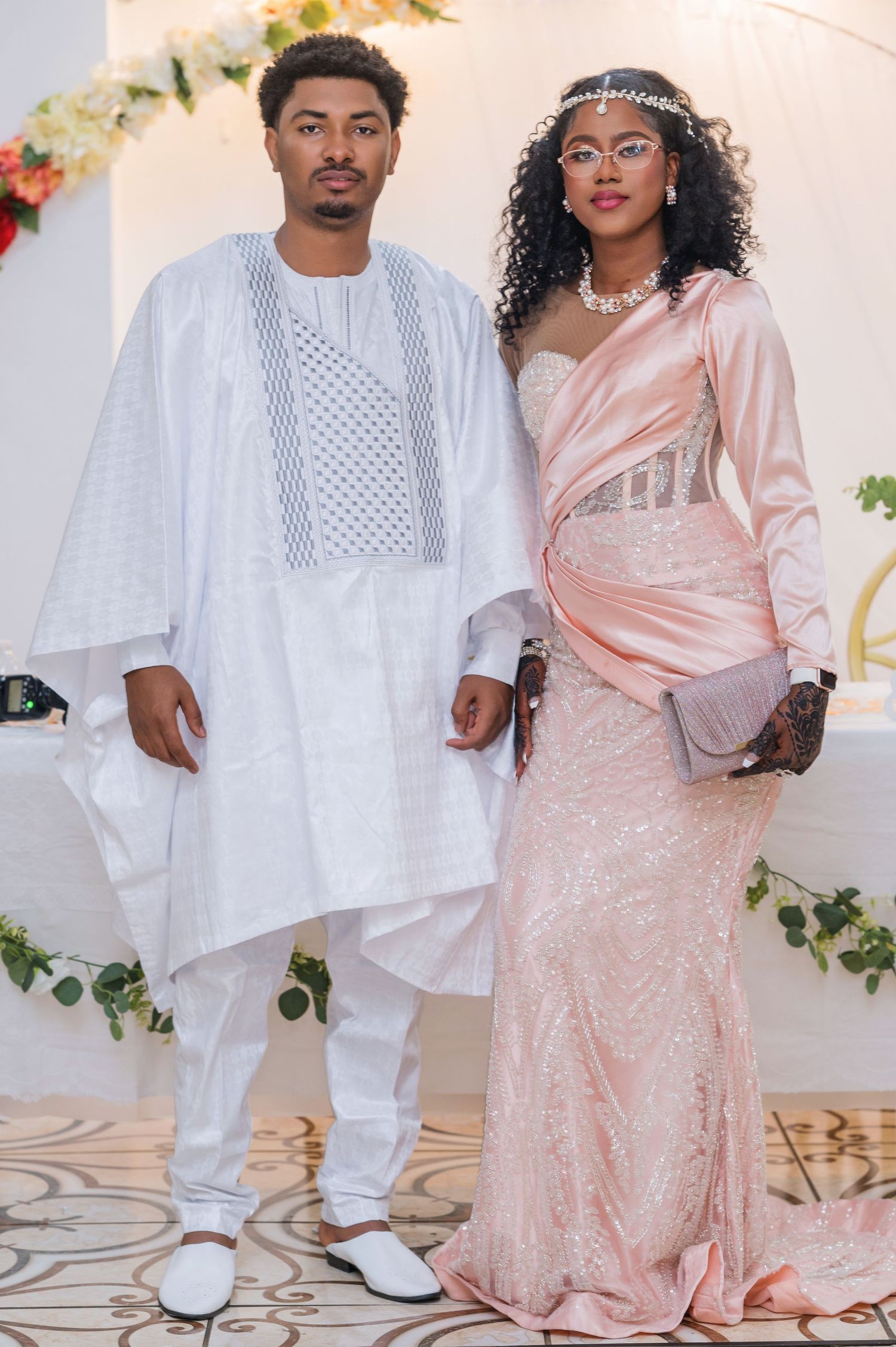 A couple poses in elegant traditional attire, with him in white agbada and her in a blush pink gown with silver accessories.