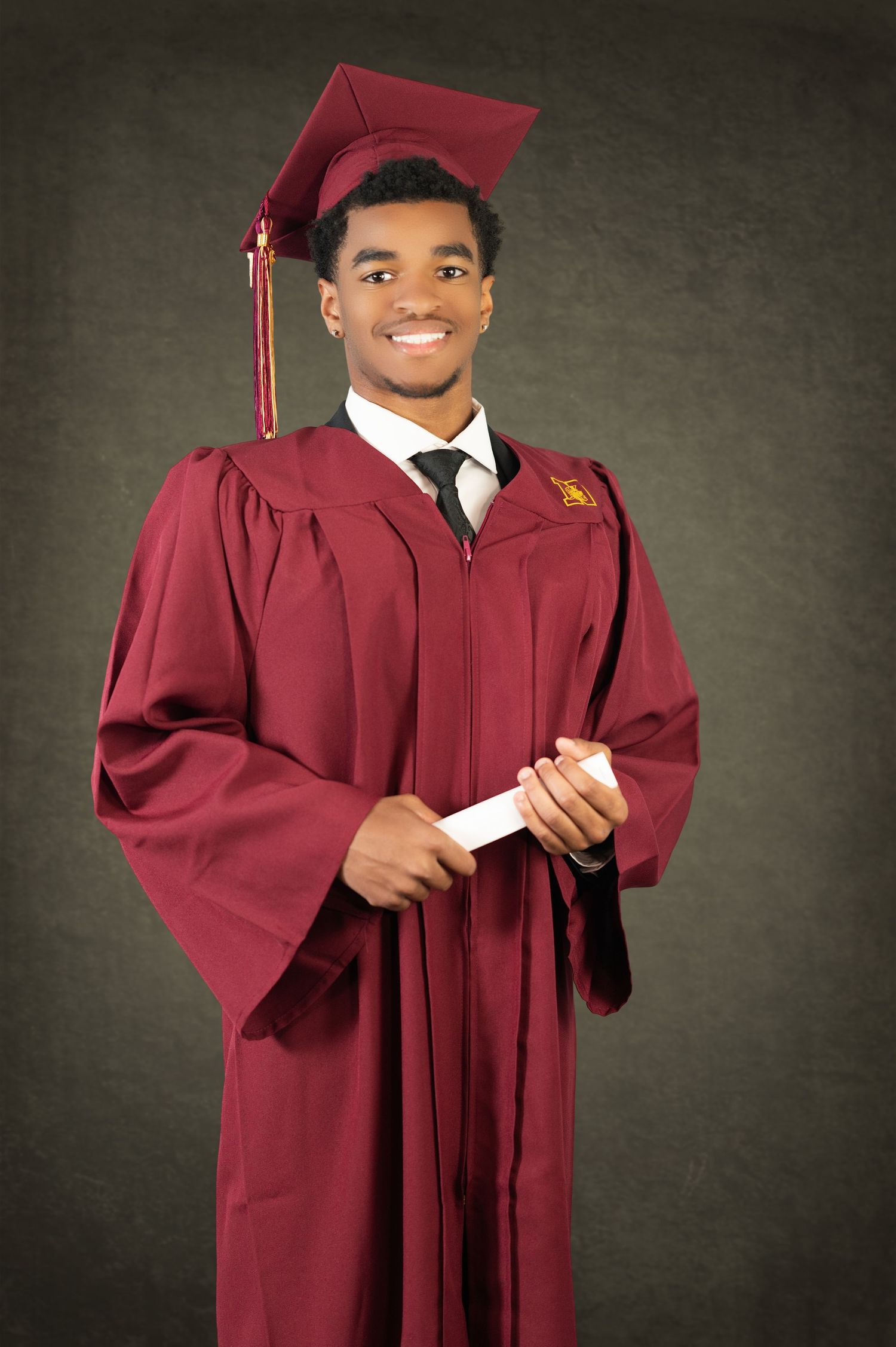 A graduate in maroon cap and gown holding a diploma while smiling against a dark background.