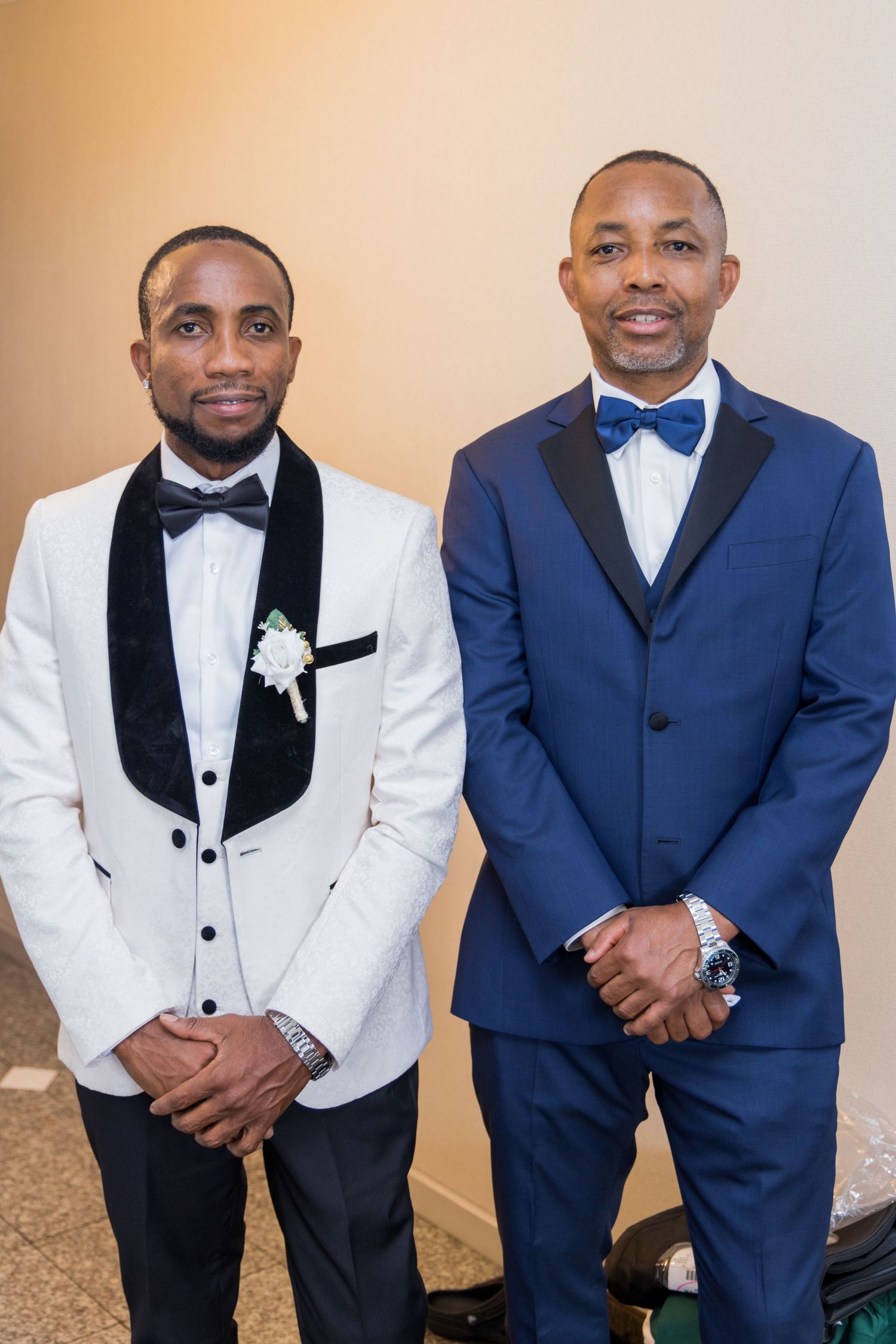 Two men in formal attire pose together, one wearing a white tuxedo jacket and the other in a navy blue suit.