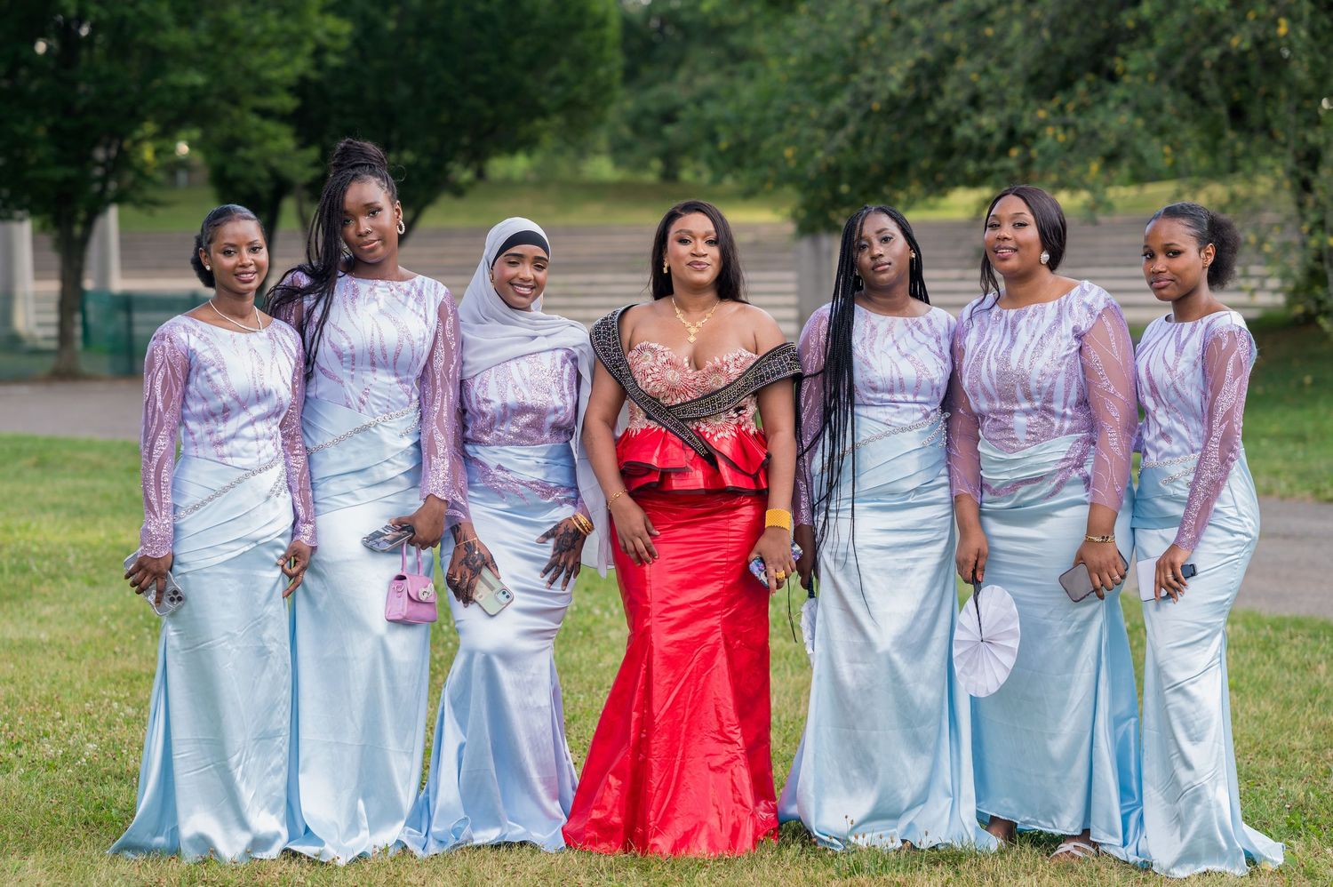 A group of women in elegant light blue bridesmaid dresses posed with someone in a striking red formal gown outdoors.