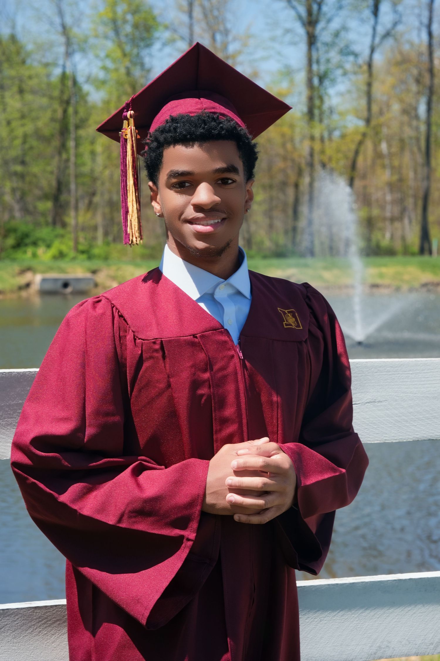 A graduate in a burgundy cap and gown poses outdoors on a sunny day with a water fountain in the background.