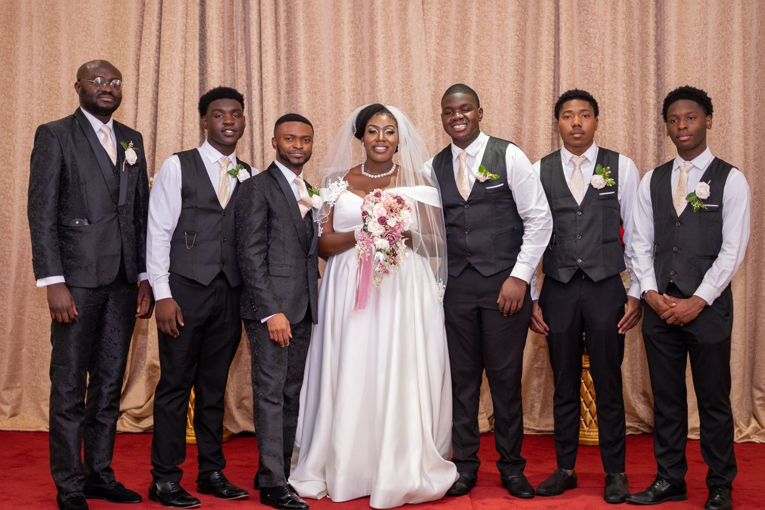 Formal wedding party wearing white and black attire poses together against a beige backdrop with red carpet.
