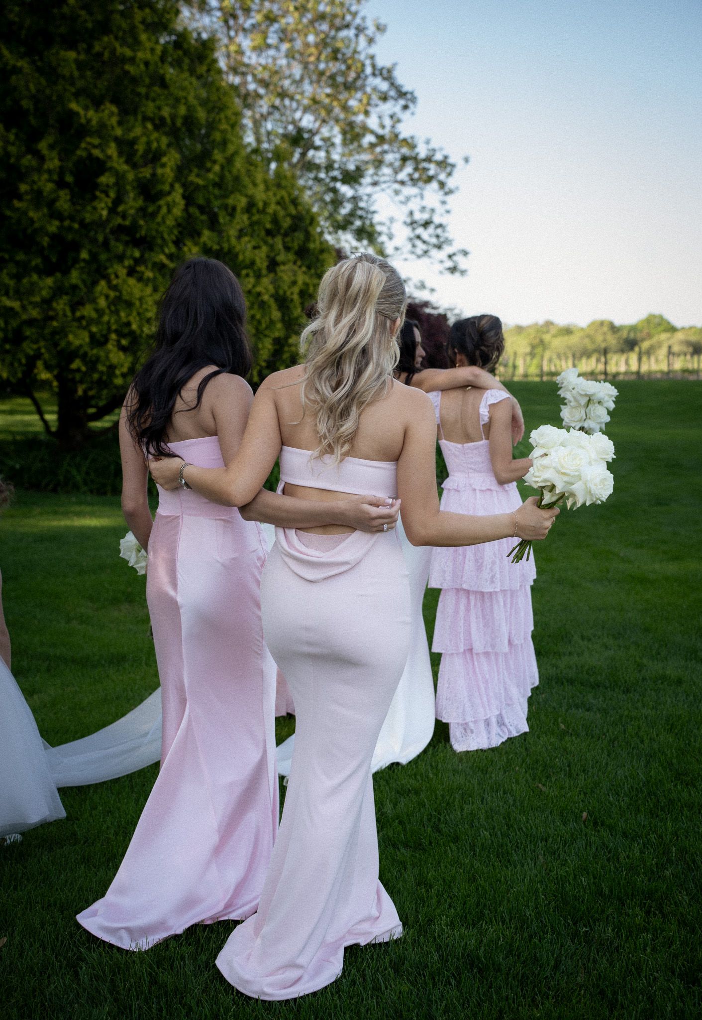 Bridesmaids in soft pink dresses walking arm in arm with the bride across a manicured lawn at a Hamptons estate wedding.