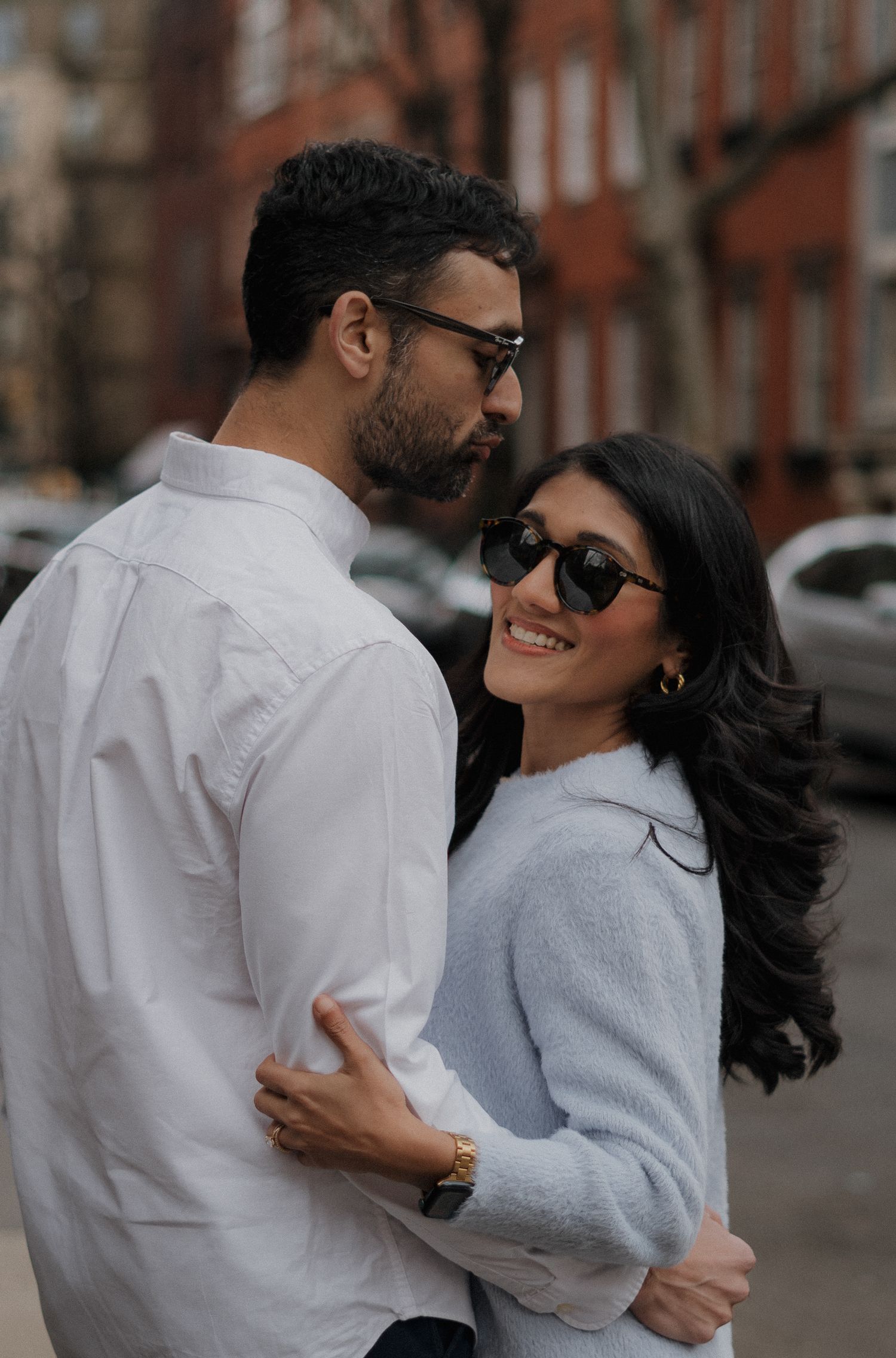Well-dressed couple in sunglasses share an intimate embrace on a city street with brick buildings in the background.