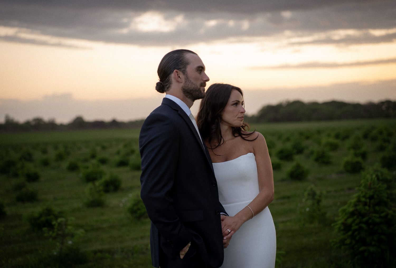 Bride and groom embracing at golden hour in an open Hamptons vineyard, gazing toward the horizon
