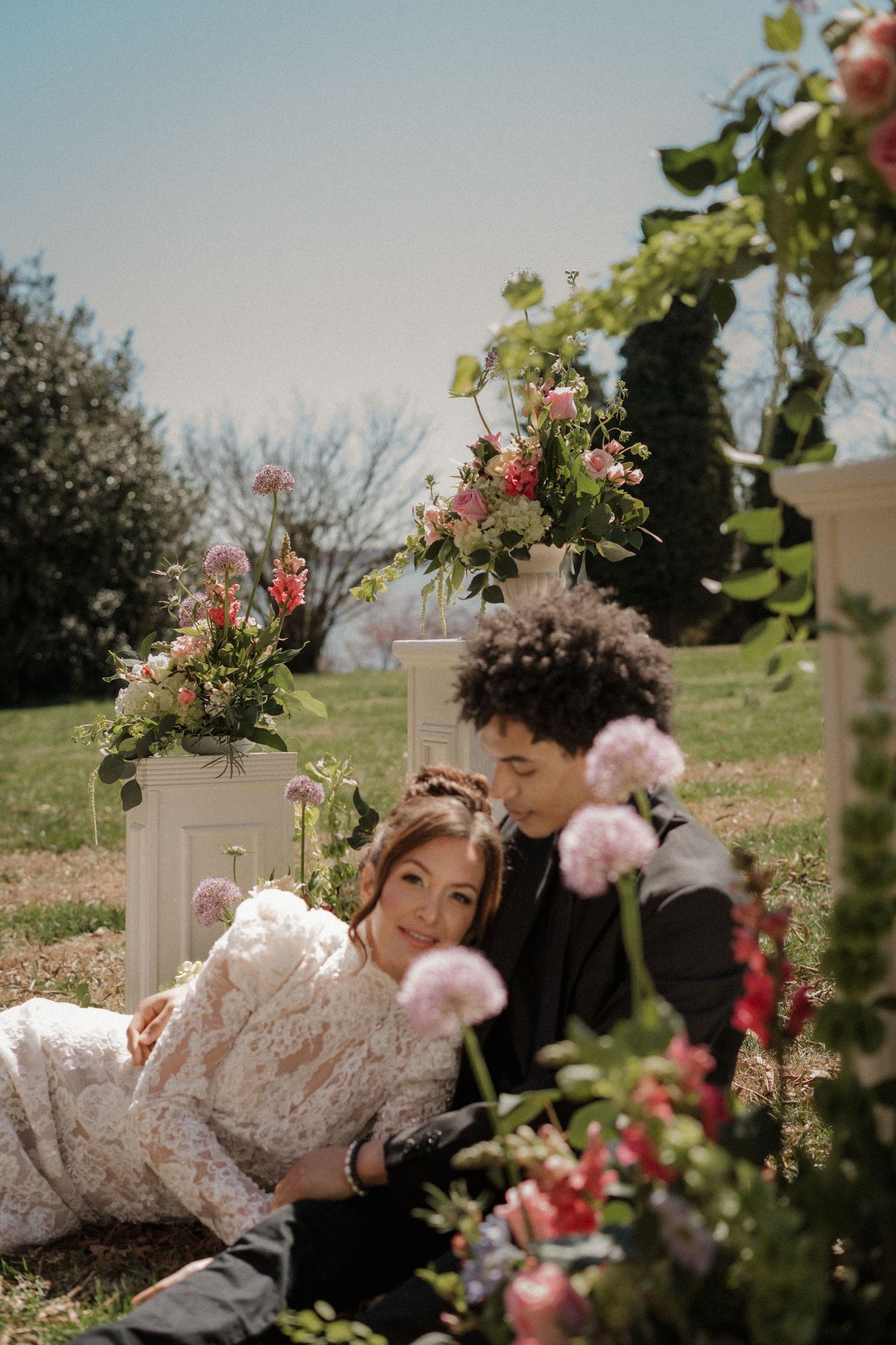 A couple rests among elegant floral arrangements and pink roses in a romantic garden setting during a wedding photoshoot.