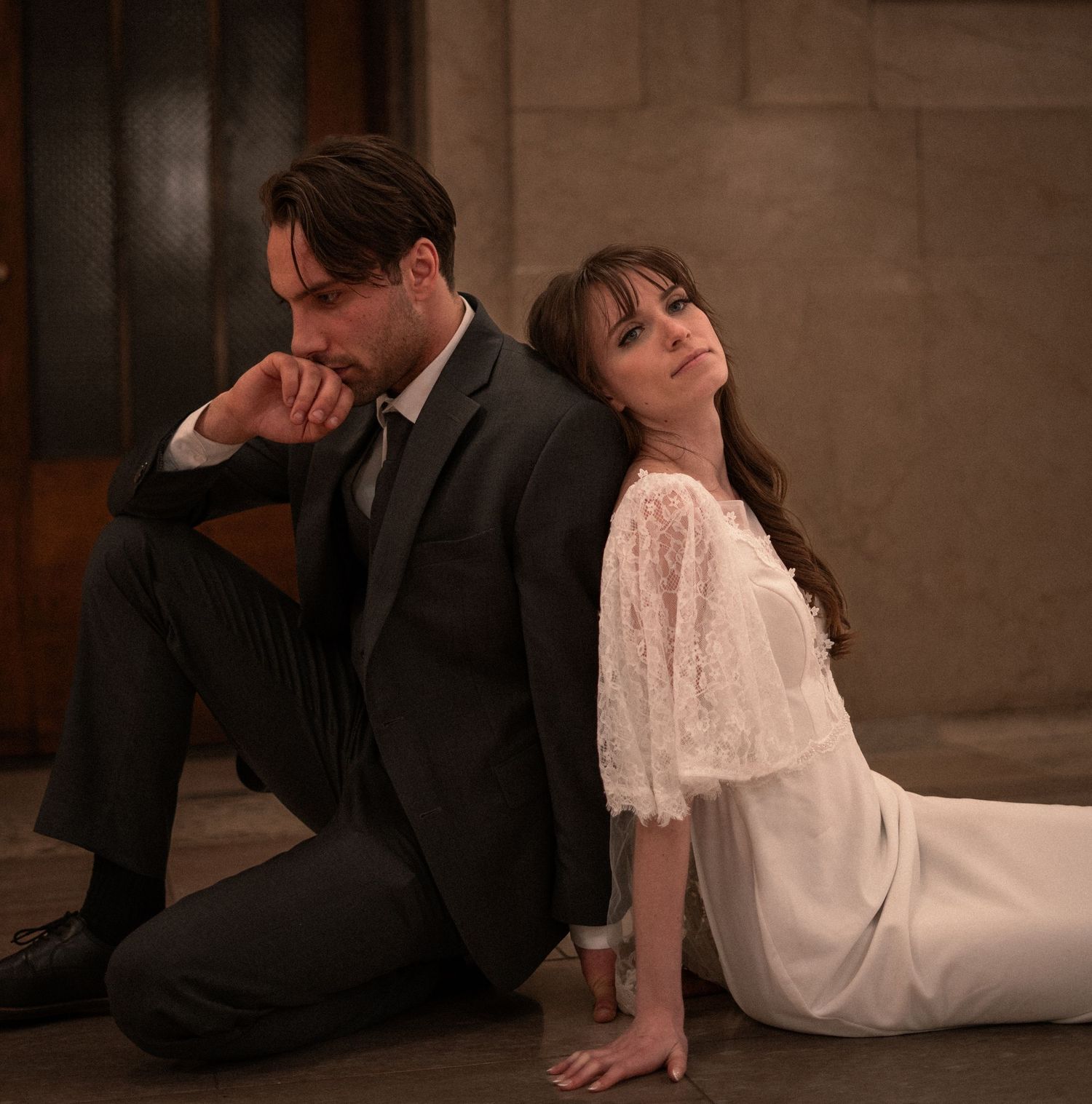 A couple in formal wear sit back to back in moody lighting, with him in a suit and her in a white lacy dress.