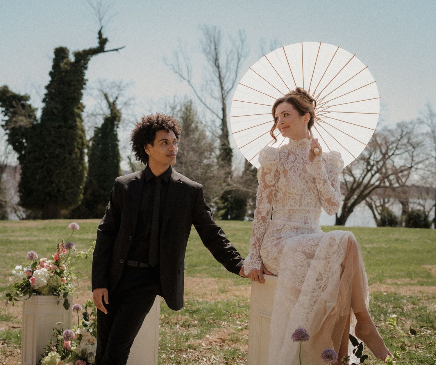 A couple in elegant attire walk in a garden, with the person in white holding a delicate parasol against the sunlight.