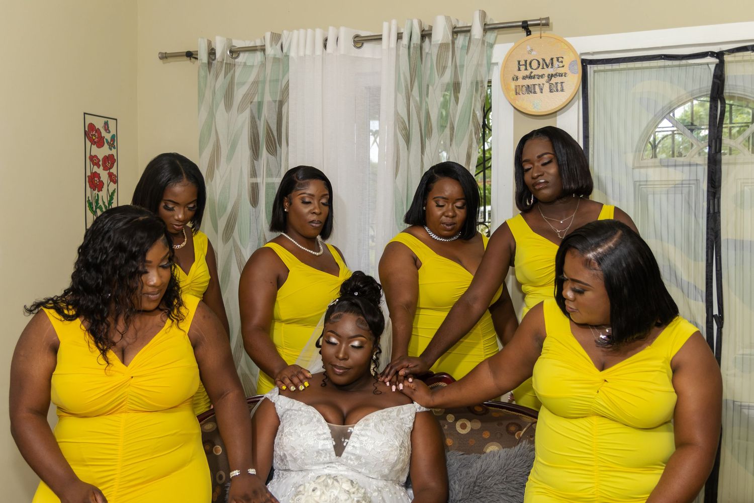 Bride in a white wedding gown sitting on a couch, surrounded by six bridesmaids dressed in vibrant yellow gowns, with hands placed on her.