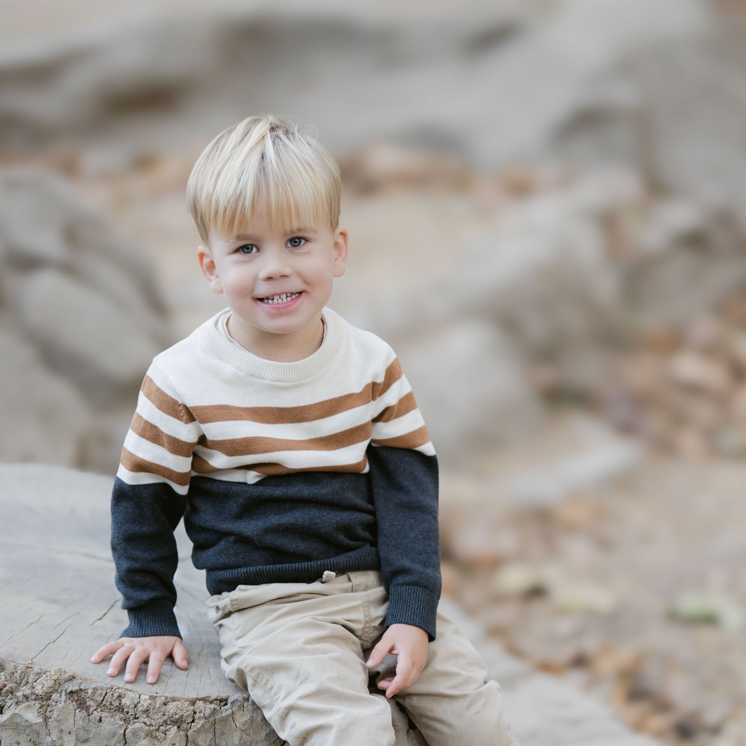 A young child in a striped sweater sits on a rocky surface outdoors while smiling at the camera.