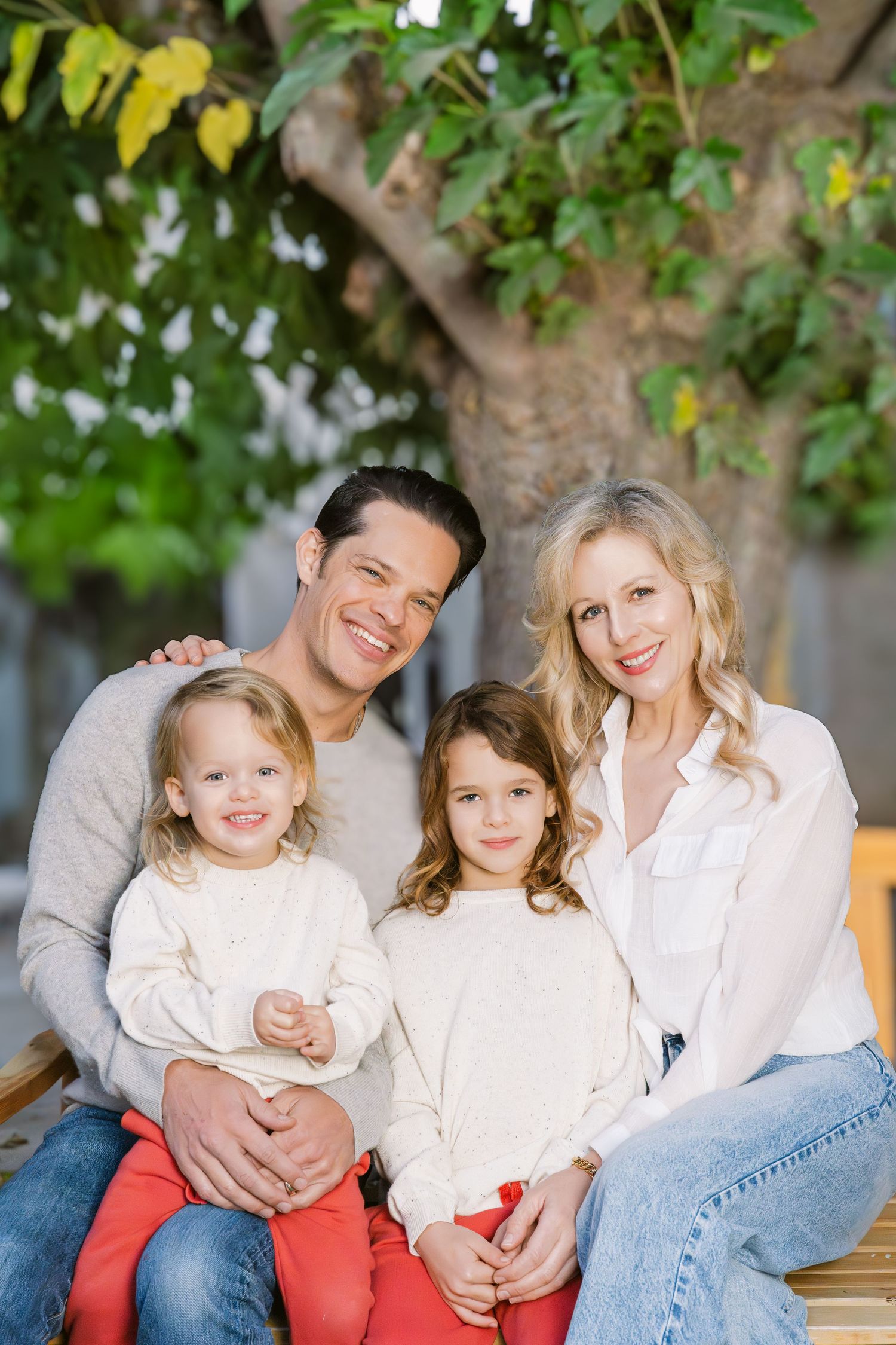A family wearing coordinated white and coral outfits sits together on a bench outdoors with green foliage in background.