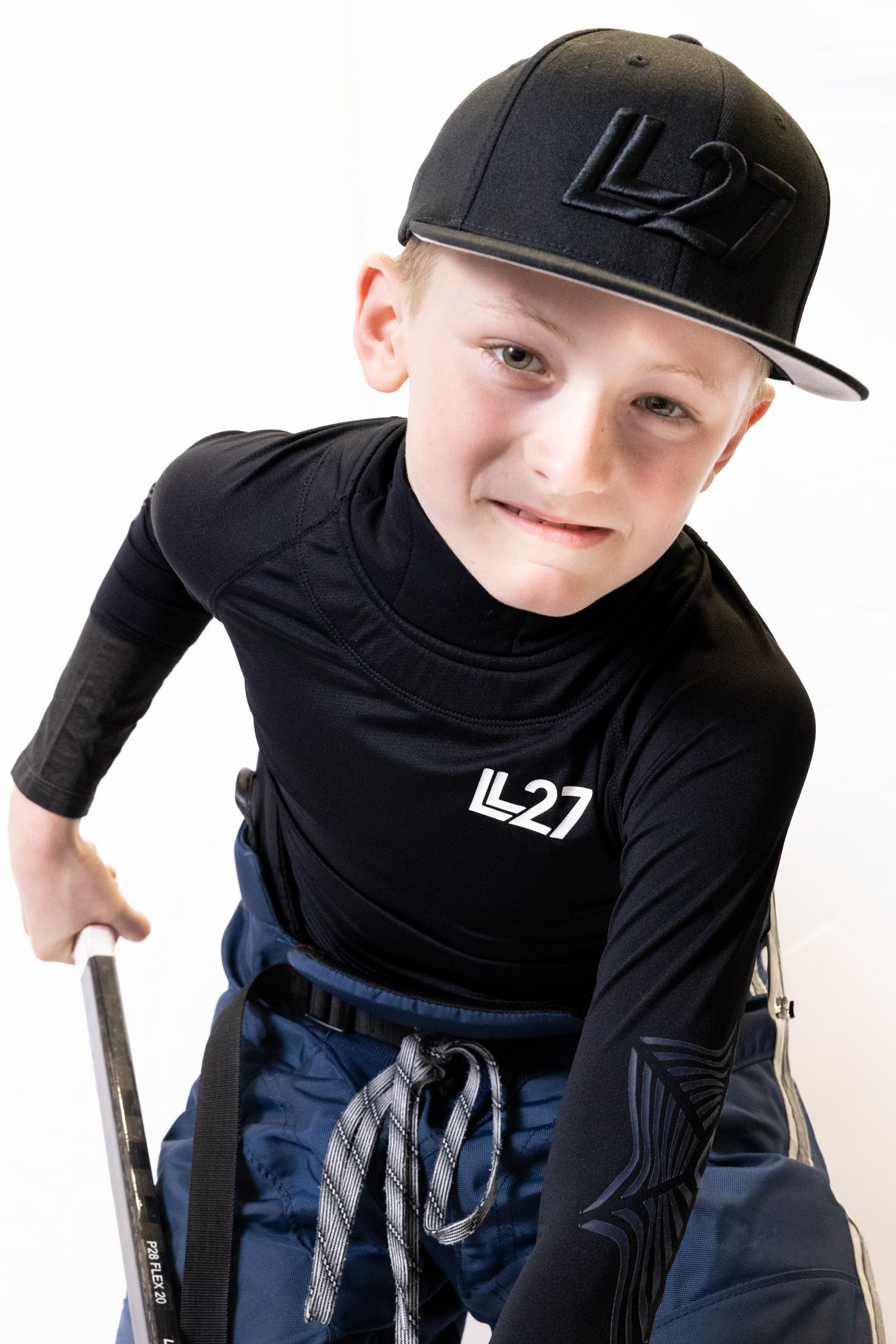 Young boy in black LL27 hockey gear, wearing matching cap, holding a stick, smiling at camera.