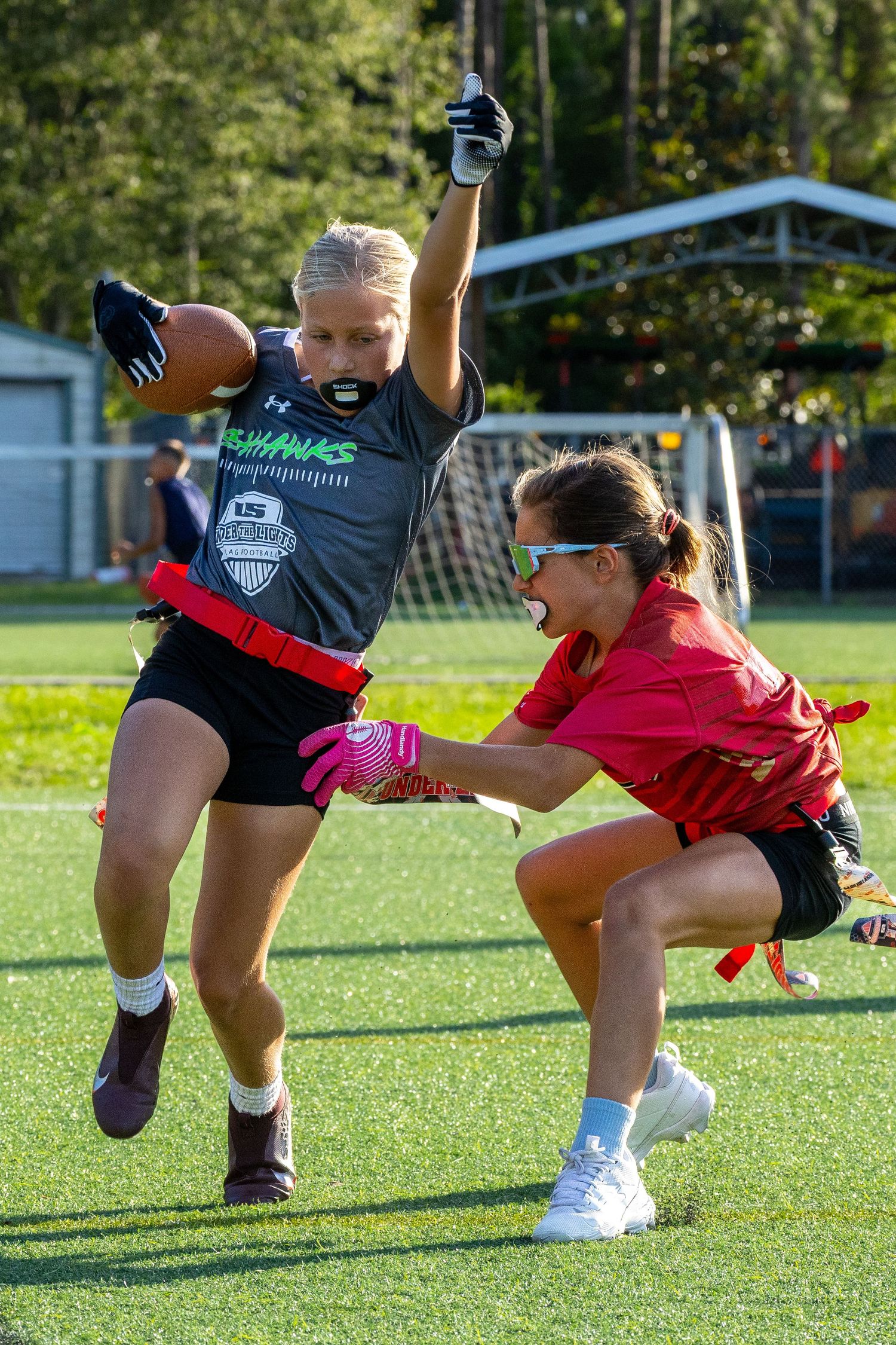 Two women compete in a flag football game, one carrying the ball while the other reaches for her flag on a turf field.