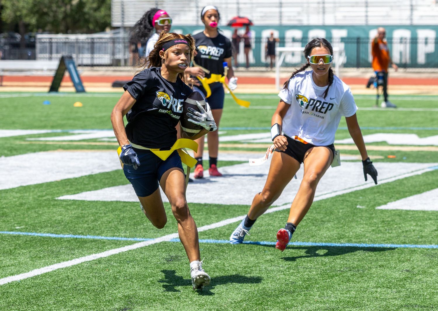 Two girls compete in a flag football game on a green turf field during a sunny outdoor youth sports event.