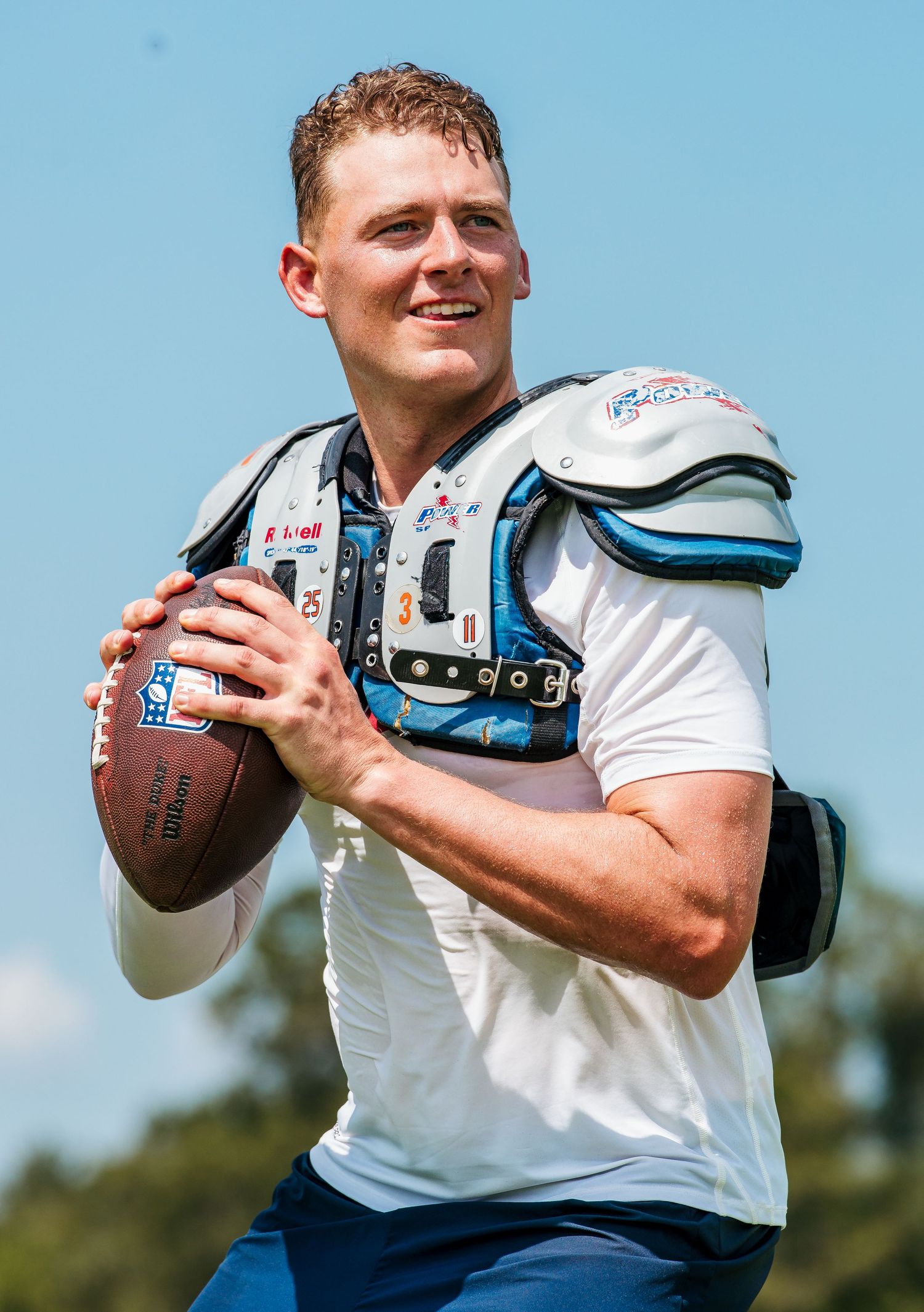 Football player in white jersey and shoulder pads holds a Wilson football, smiling during an outdoor practice session.