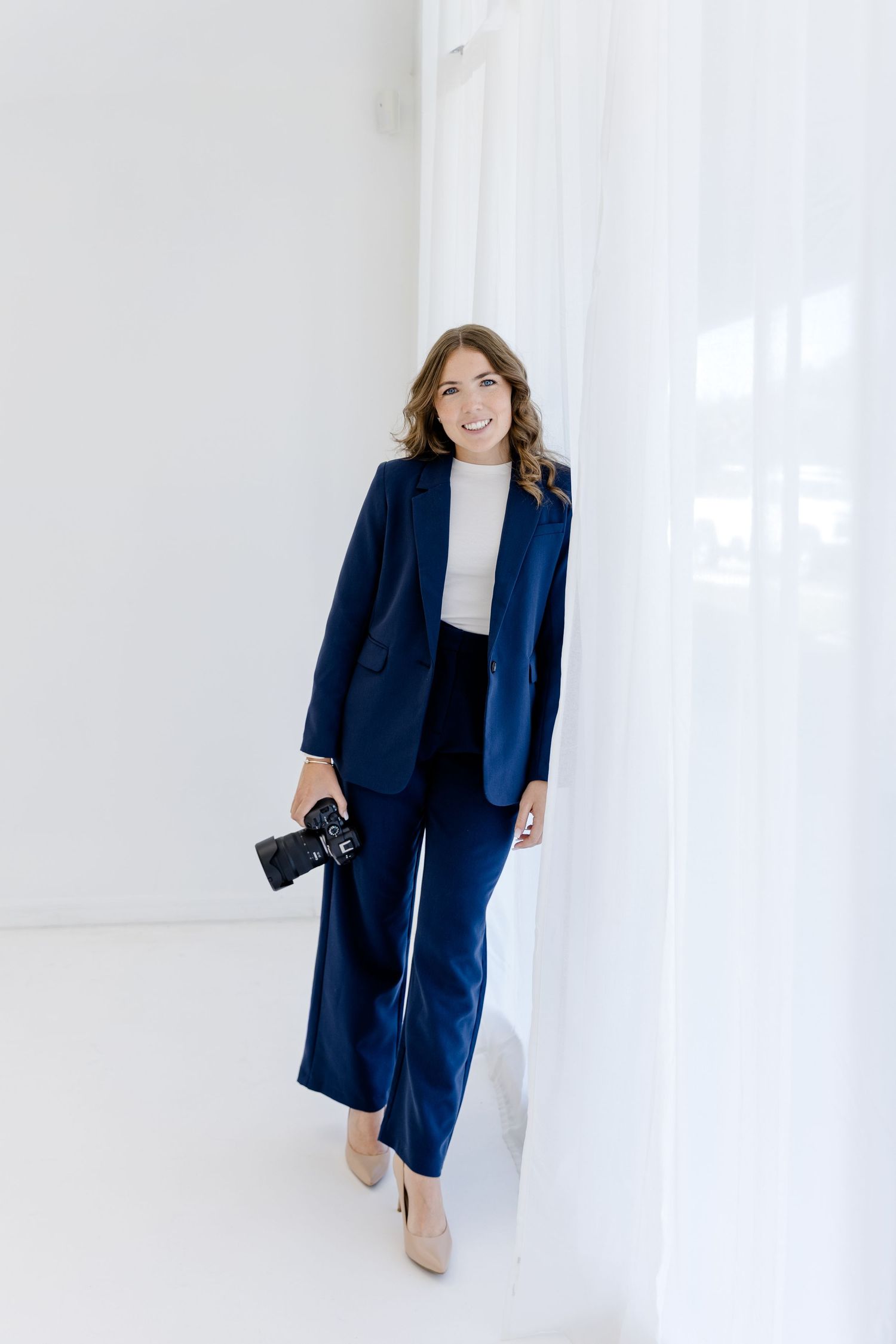 A smiling woman in a navy blue suit holds a camera while posing in a bright white studio setting.