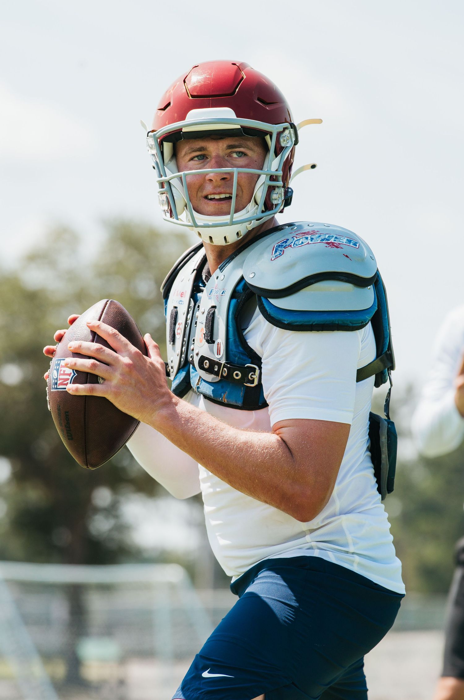 Football player in white uniform and red helmet prepares to throw a pass during an outdoor practice session.