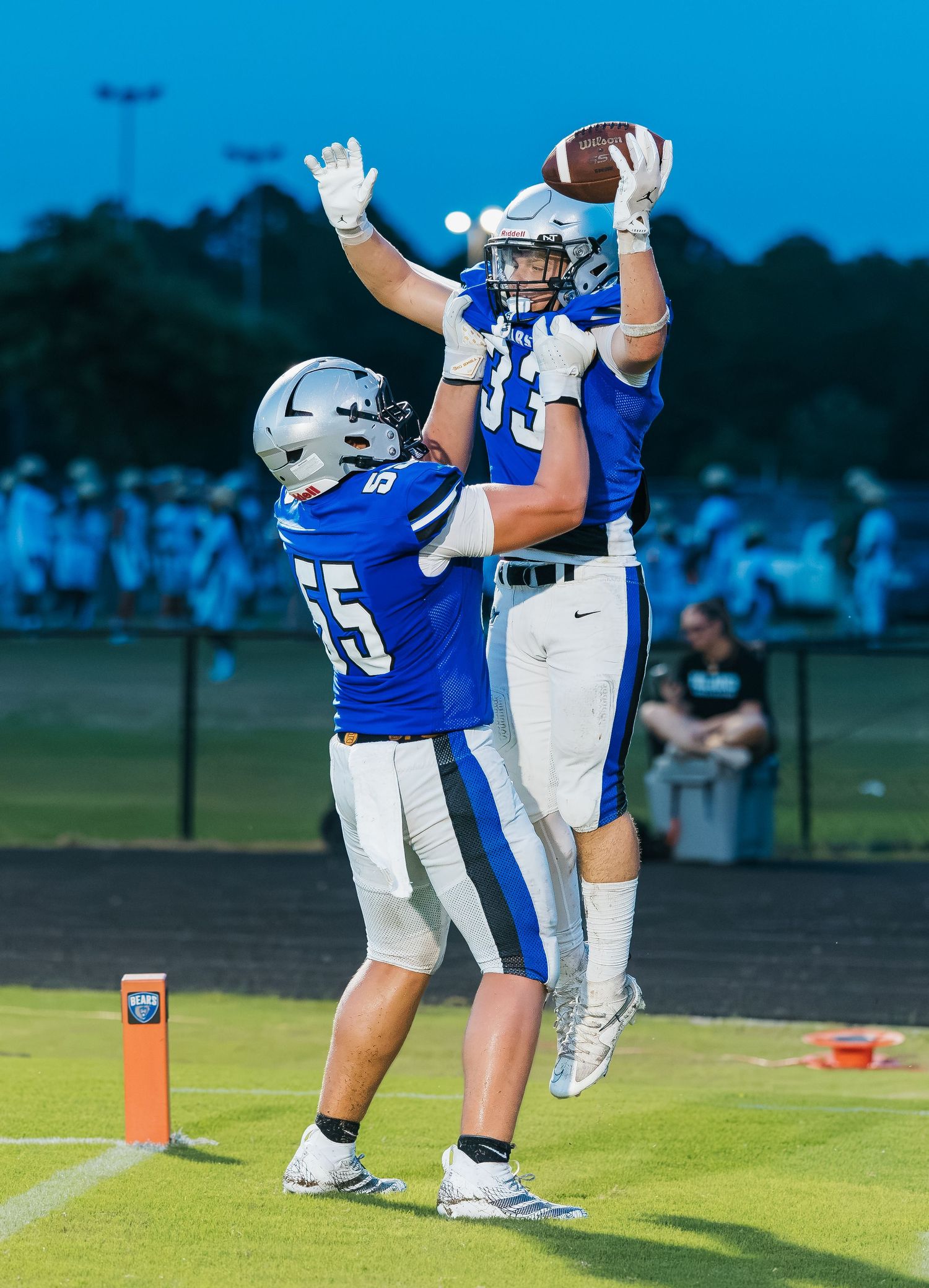 Two football players in blue uniforms celebrate a touchdown catch under stadium lights during a Friday night game.