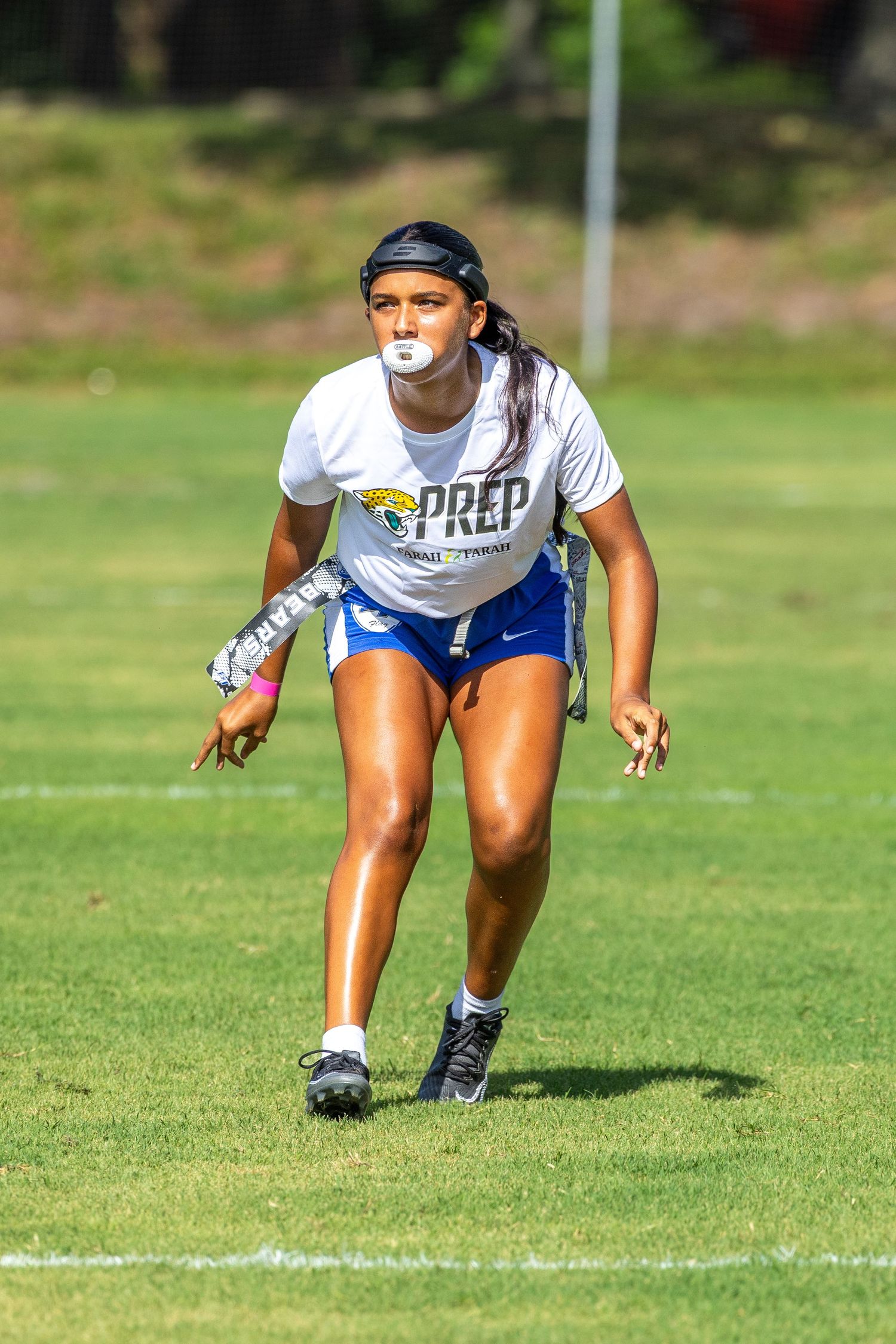 Female athlete in PREP jersey playing flag football on a green field, wearing a mouthguard and blue shorts.
