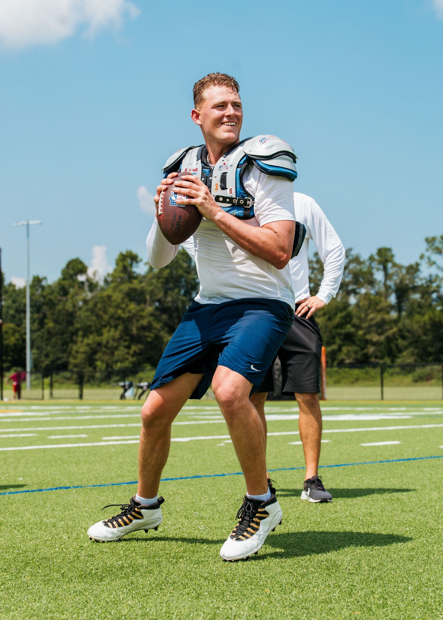 Football player in white jersey and blue shorts prepares to throw a football on a green practice field on a sunny day.