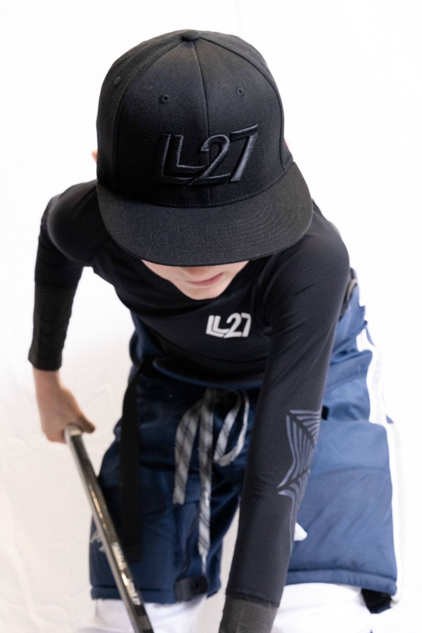 Young hockey player wearing black L27 branded cap and jersey, holding a hockey stick in a studio setting.