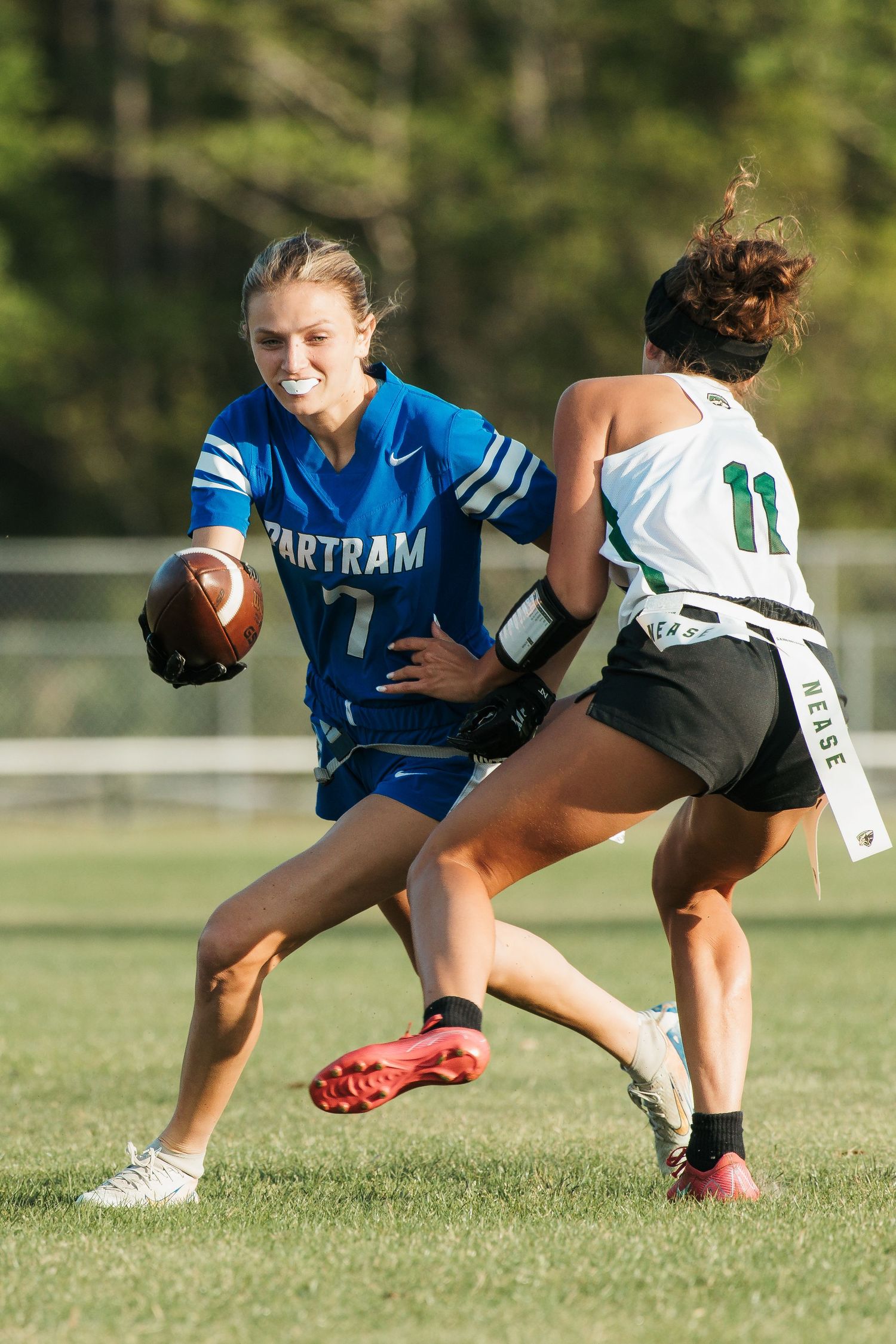 Female flag football player in blue Bartram jersey runs with ball while defender in green number 11 jersey attempts to pull flag.
