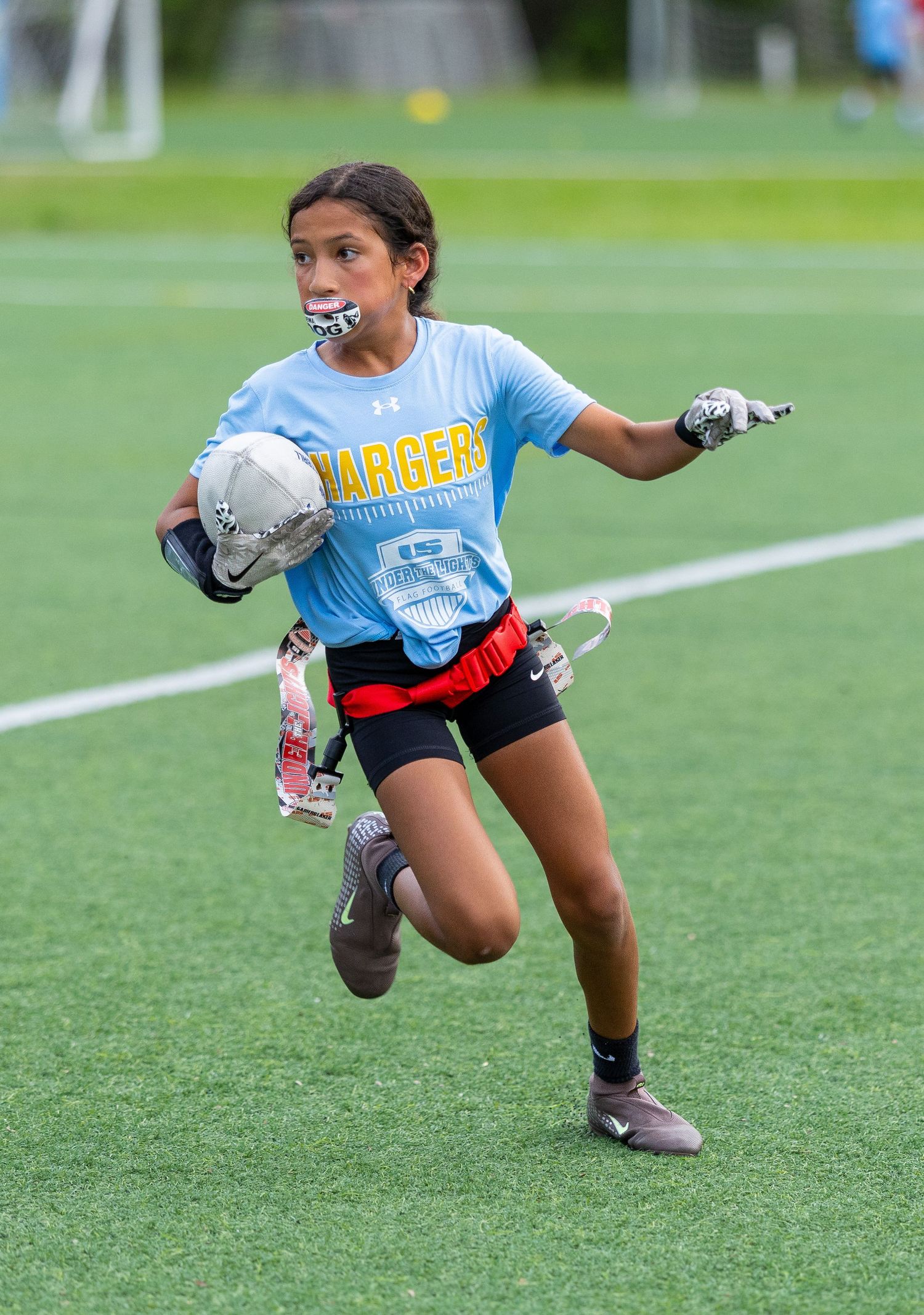 Young girl in Chargers jersey runs with football during flag football game on green turf field.