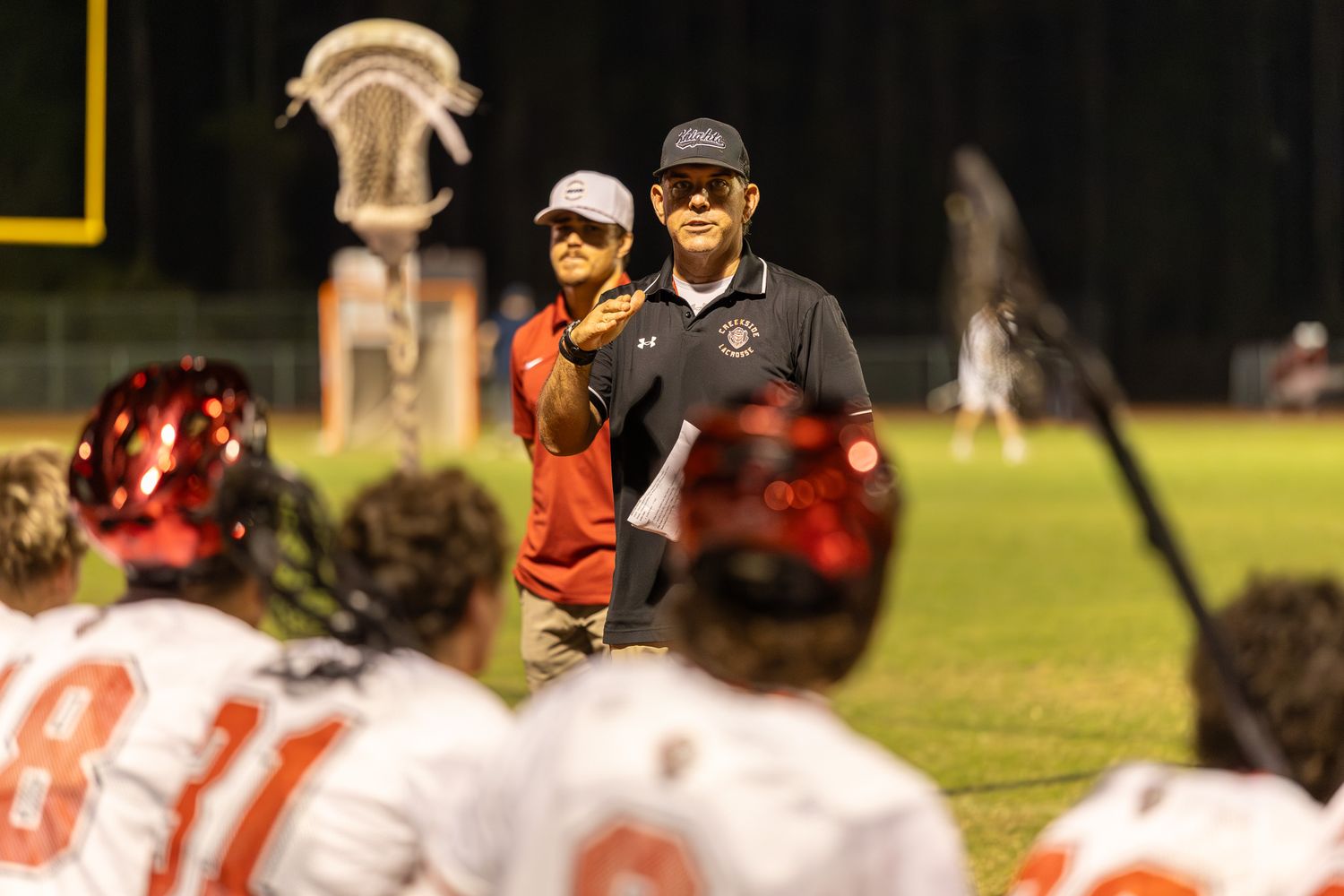 Lacrosse coach in black polo addresses players in white and red helmets on field at night game.