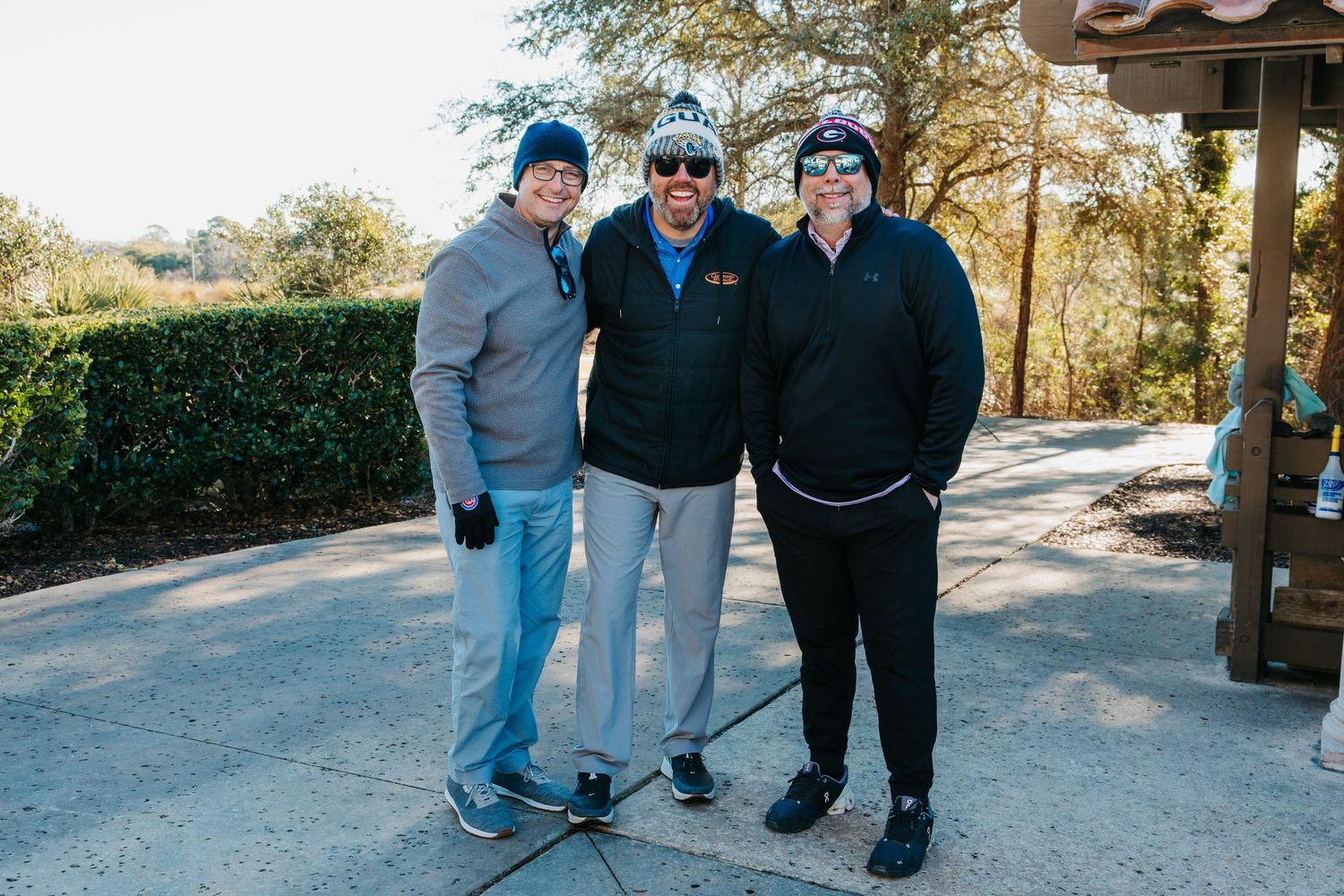 Three friends pose together outdoors on a patio, wearing winter hats and jackets on a cool day.