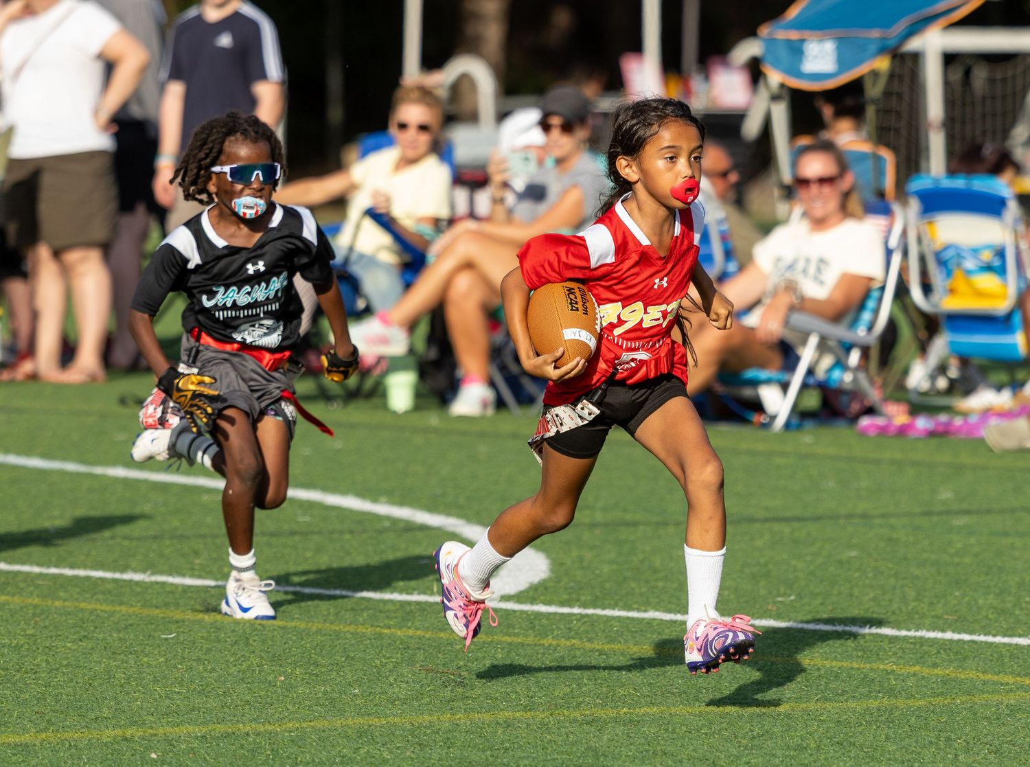 Young girls playing flag football, with a player in red uniform running with the ball during a youth sports game.