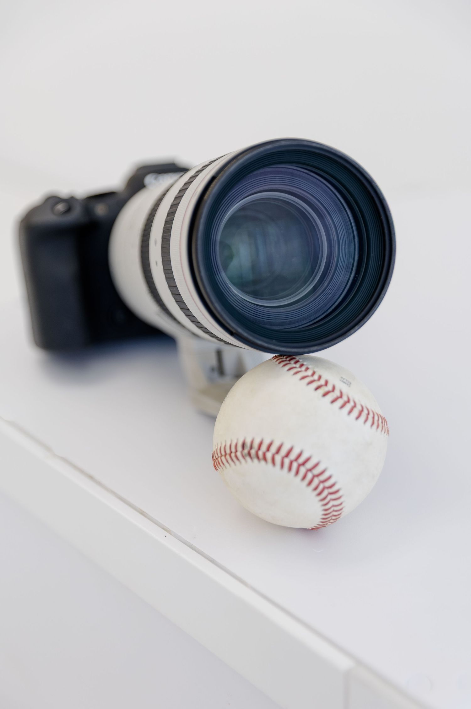A white telephoto camera lens with DSLR body rests beside a baseball on a white surface.