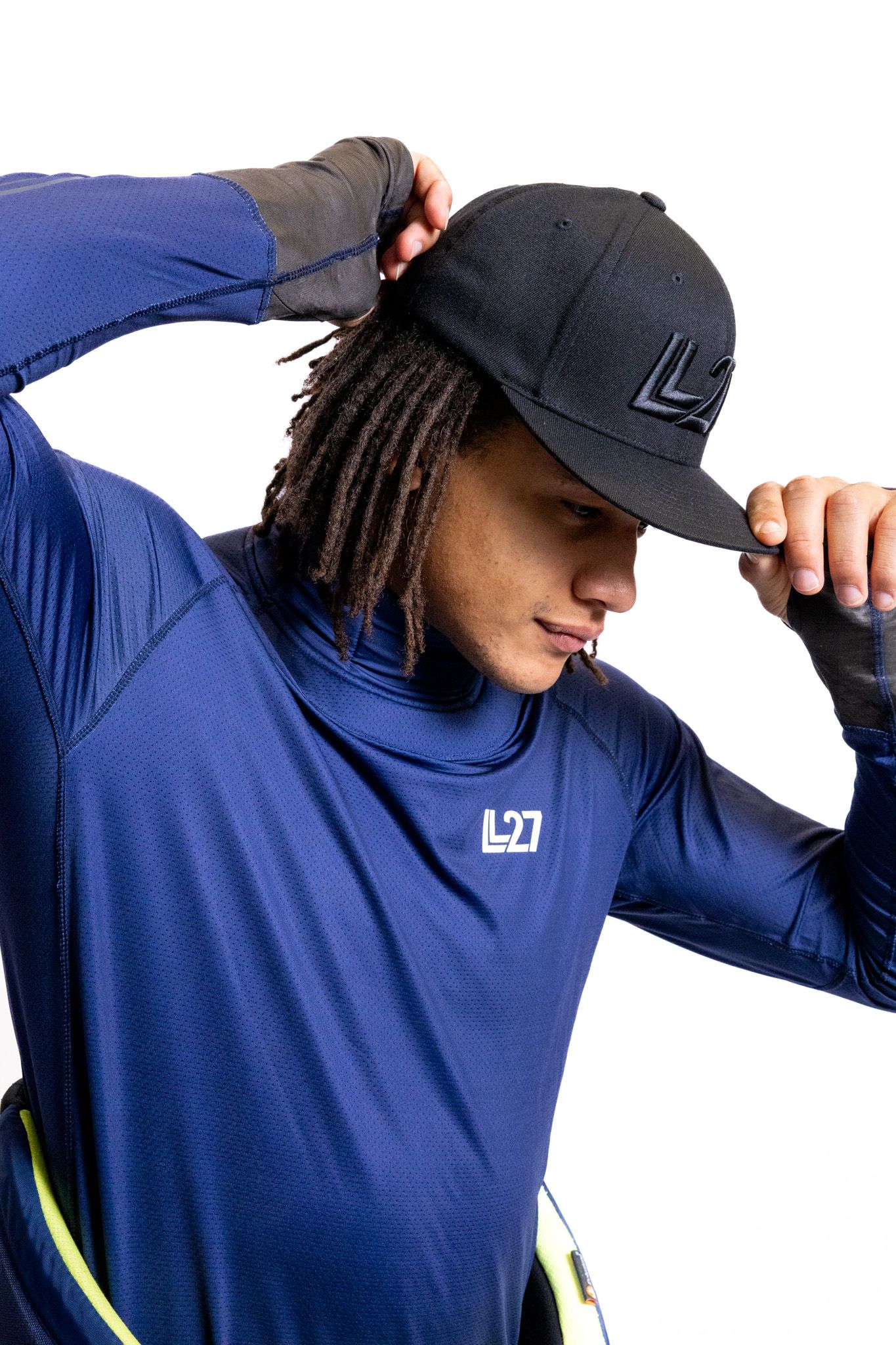 Man wearing blue long-sleeve athletic shirt and black snapback cap with logo, adjusting hat against white background.