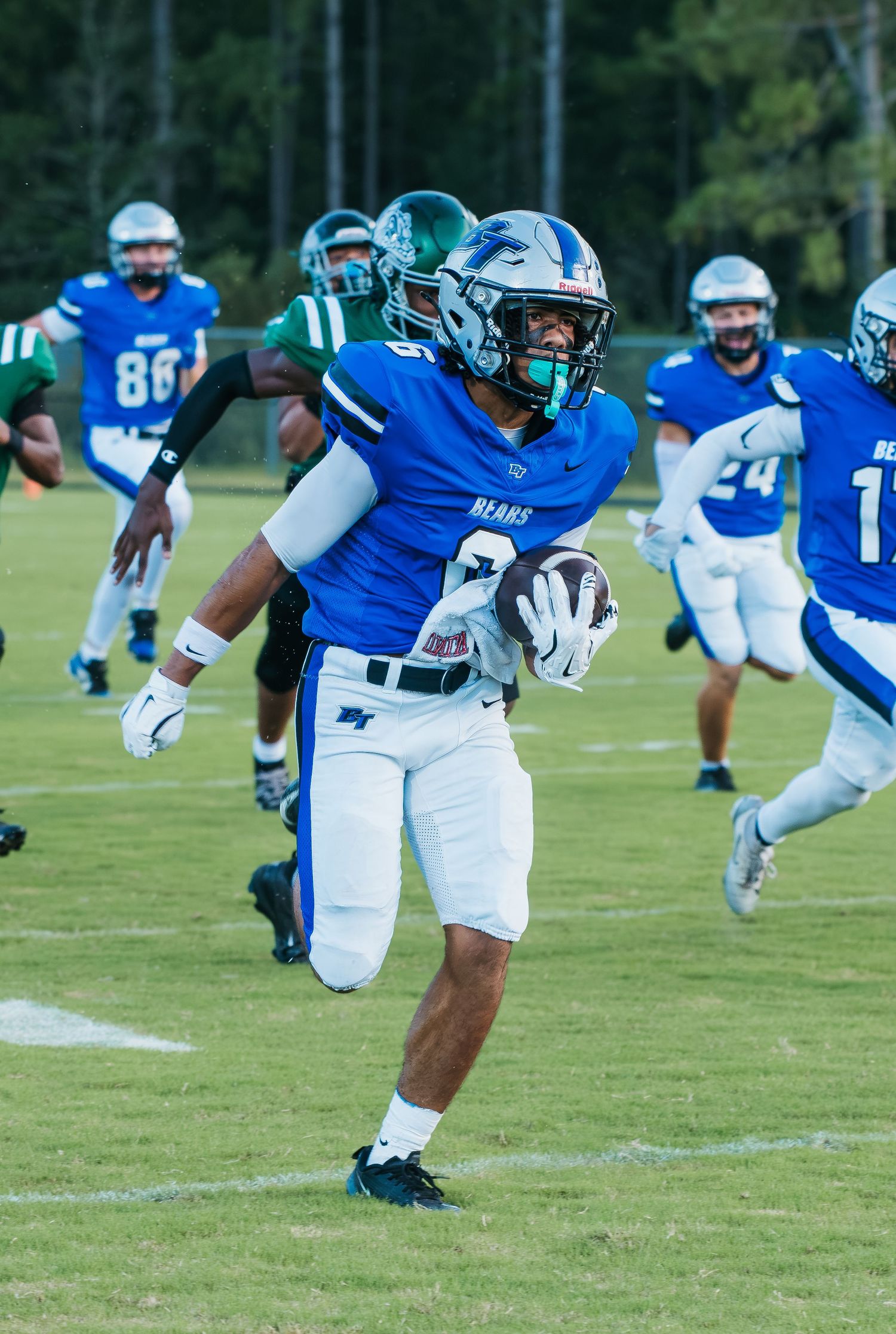 Blue team football player runs with ball during game against green team opponents on grass field.