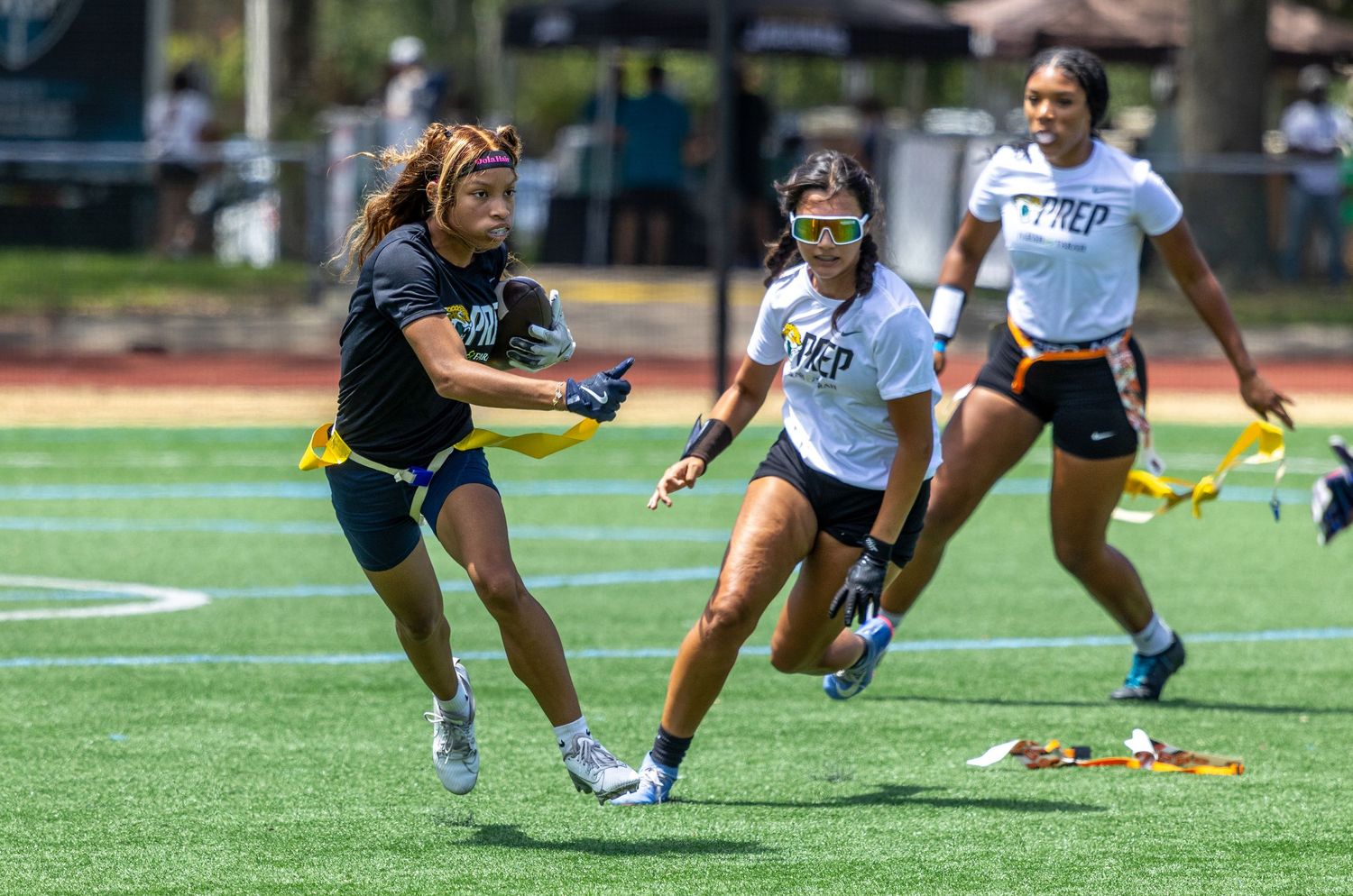 Girls playing flag football on a turf field, with one player running with the ball while defenders chase her.