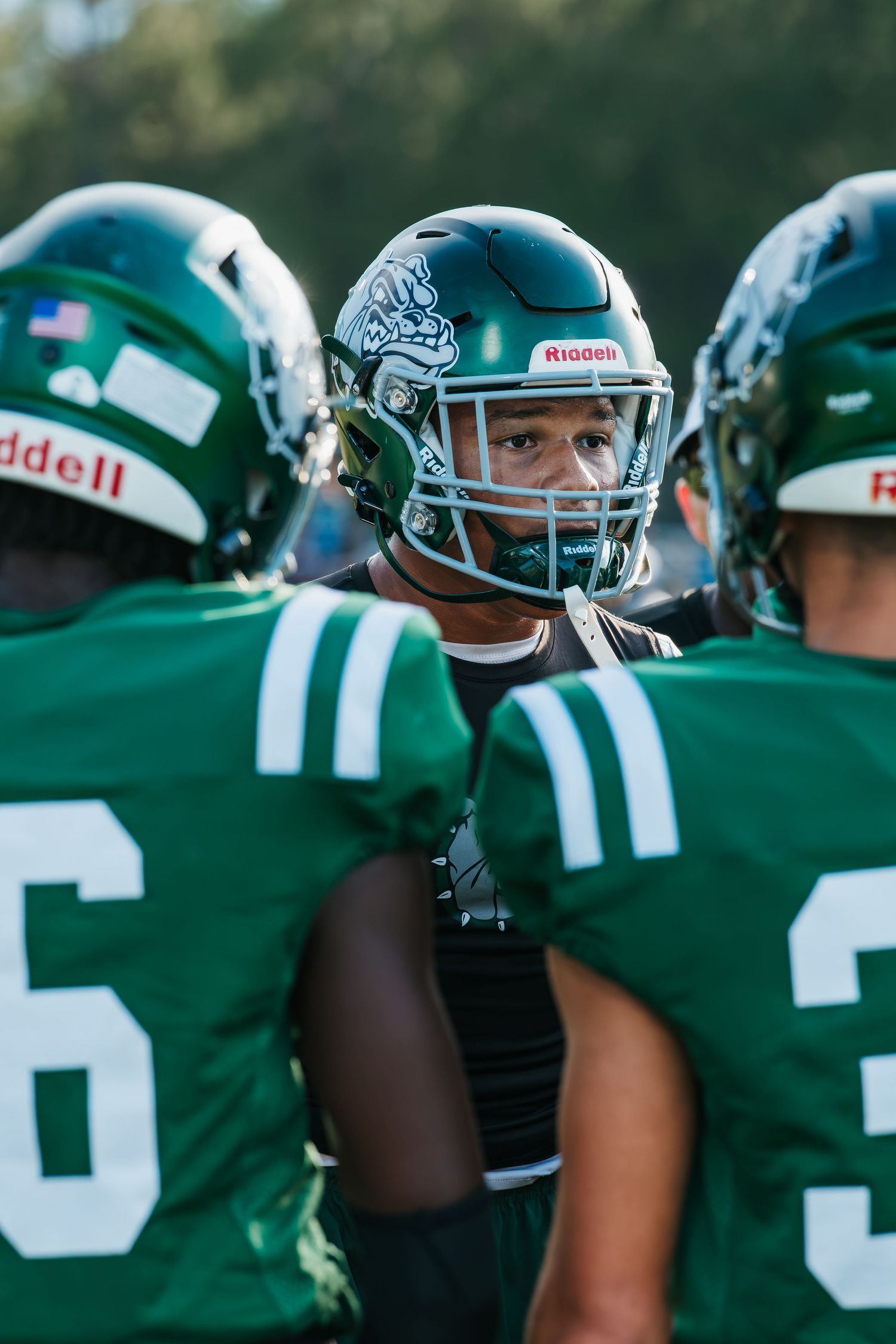 Football players in green Riddell helmets and jerseys huddle together on the field during a game.