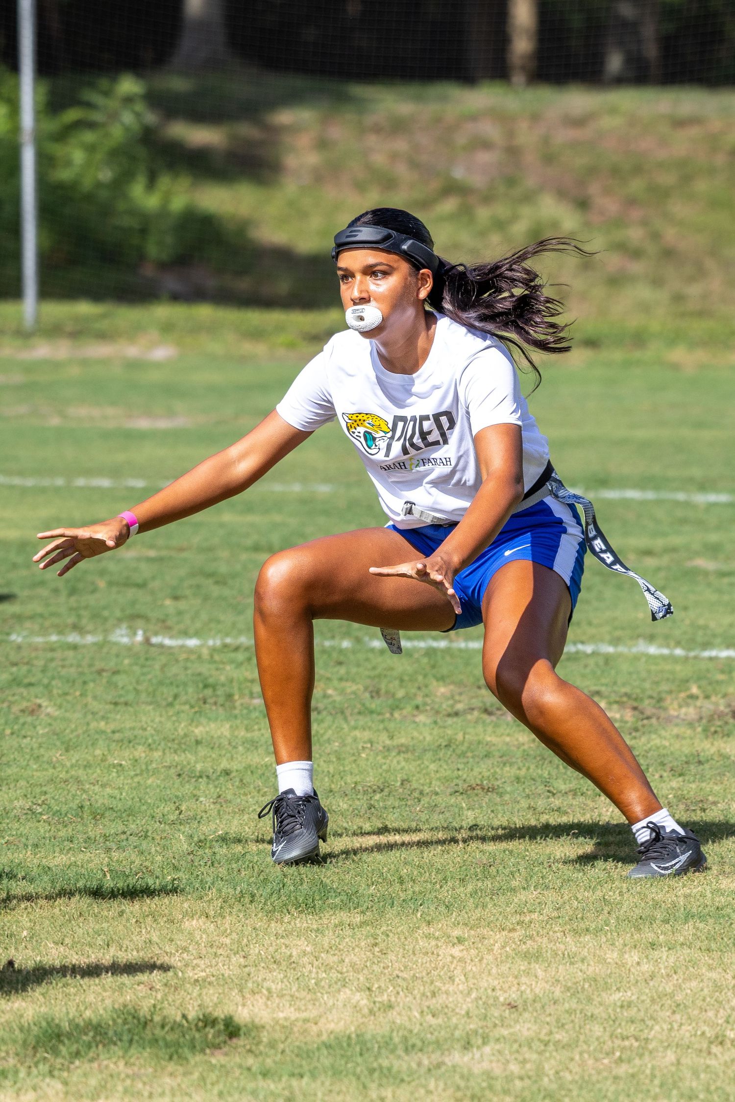 Female athlete in white PREP jersey plays flag football on grass field, mid-stride with mouthguard and blue shorts.