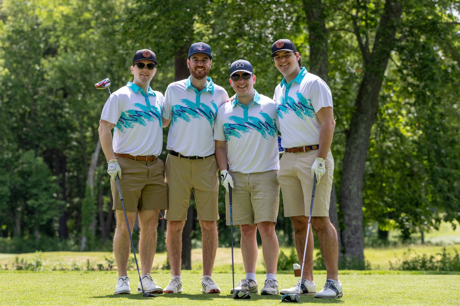 Four men in matching white and teal golf shirts and khaki shorts posing together on a sunny golf course.