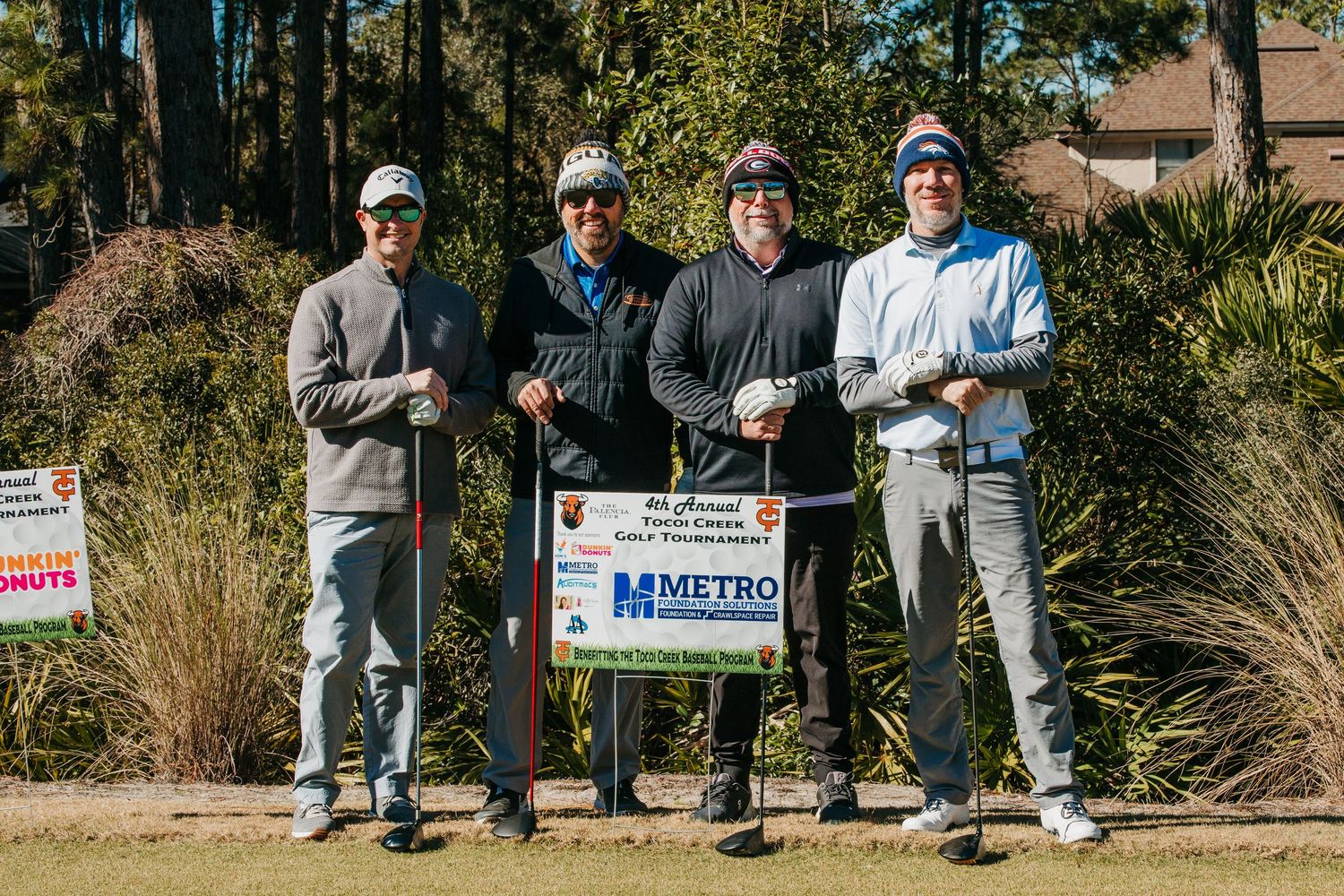 Four golfers pose together at a Metro Championship golf tournament tee box marker on a sunny day.