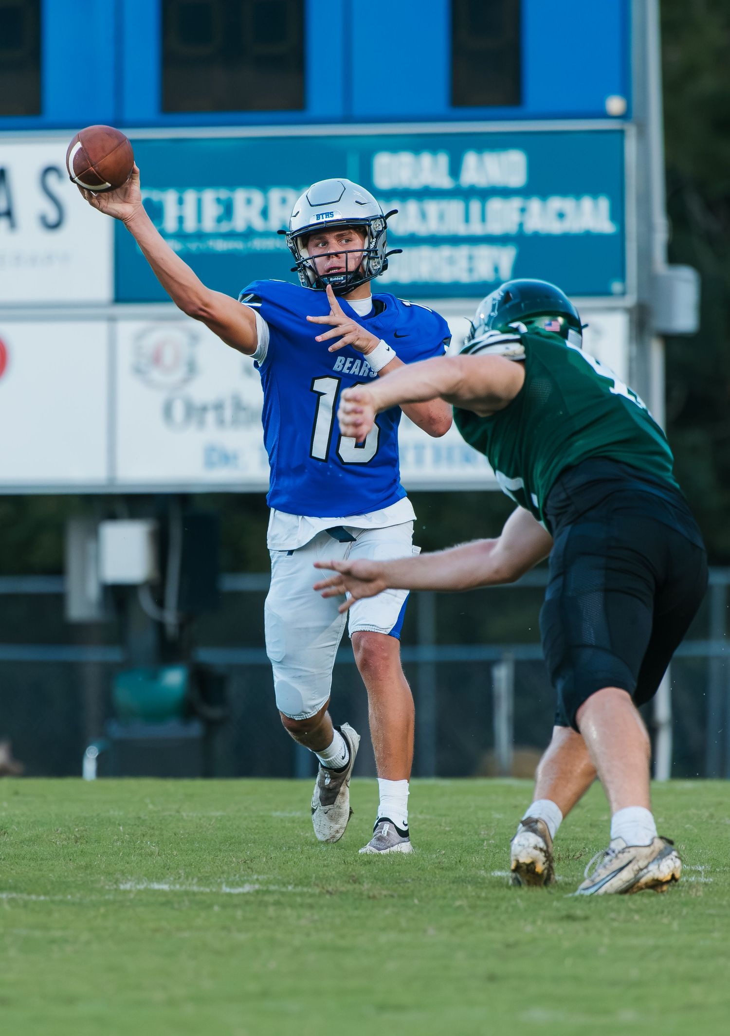 High school quarterback #10 in blue jersey throws a pass under pressure from a green-jersyed defender during a football game.