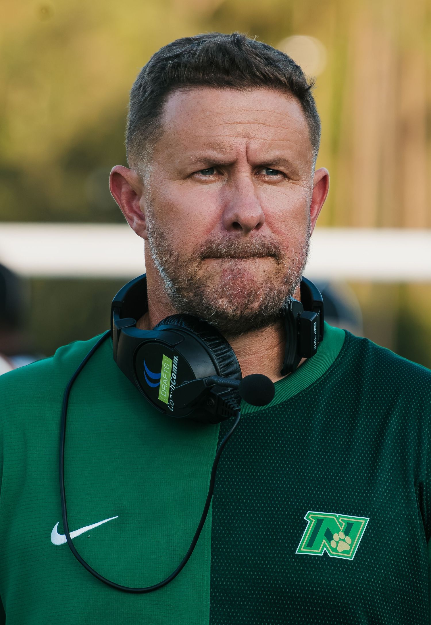Football coach in green Nike jersey with headset around neck stands on sideline with focused expression.