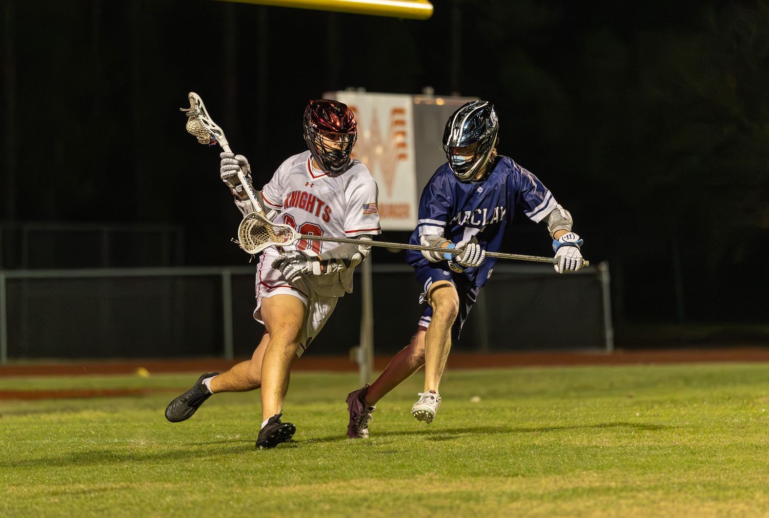 Two lacrosse players compete on a lit field at night, one in white and one in blue uniform.