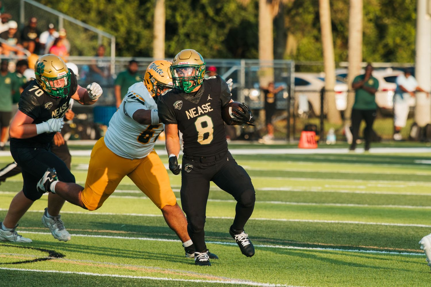 Nease High School football player #8 runs with the ball during a competitive evening game on a green turf field.