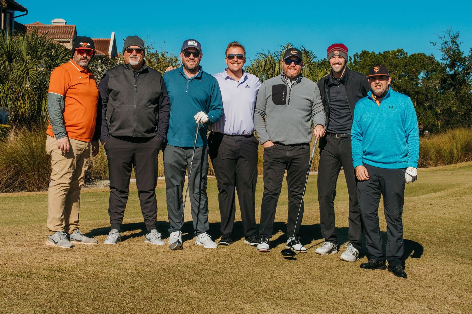 Seven men pose together on a golf course on a sunny day, dressed in casual golf attire.