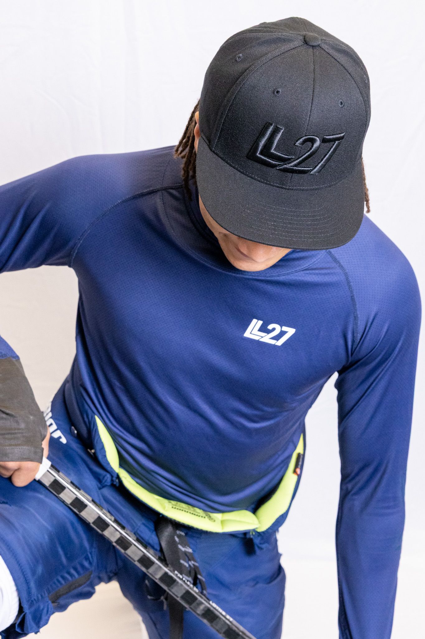 Man wearing navy LL27 long sleeve compression top and black LL27 snapback cap, holding ski equipment.