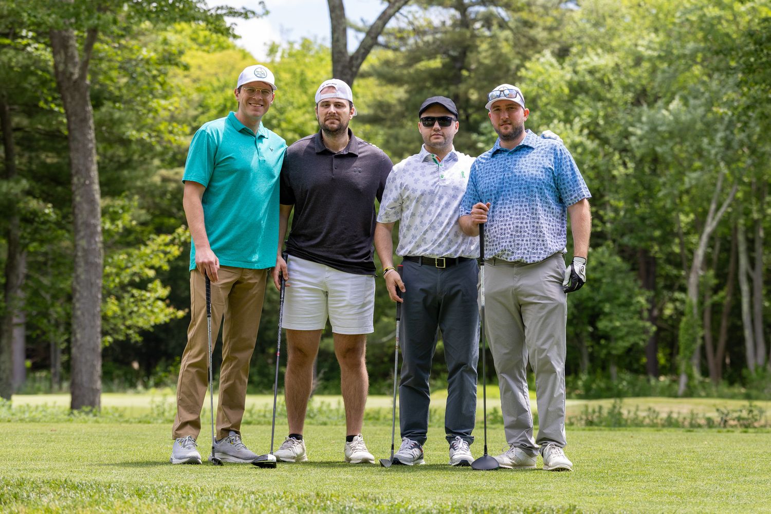 Four men posing with golf clubs on a lush green golf course on a sunny day.