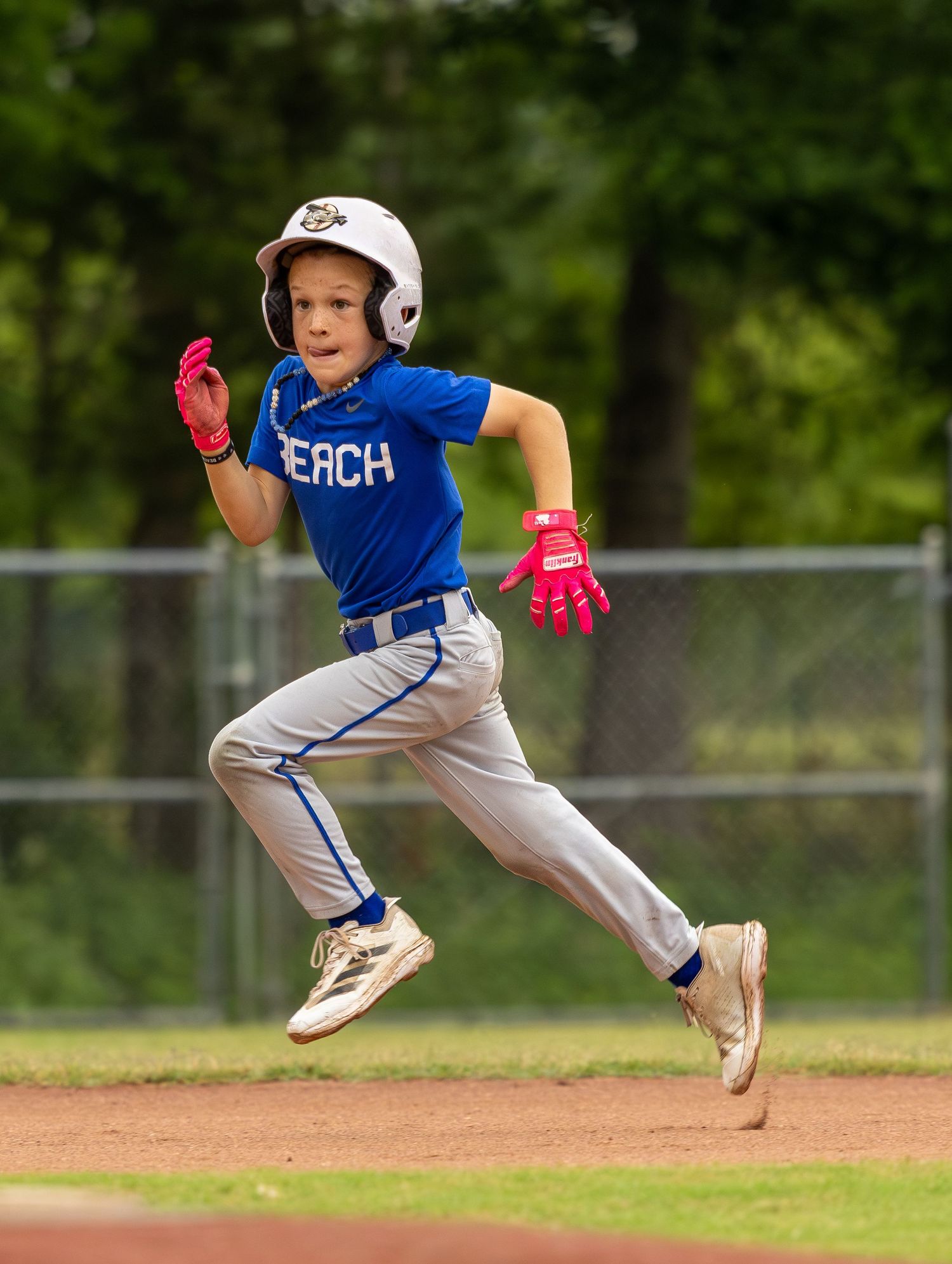 Young baseball player in blue BEACH jersey sprints energetically on a dirt field during a game.
