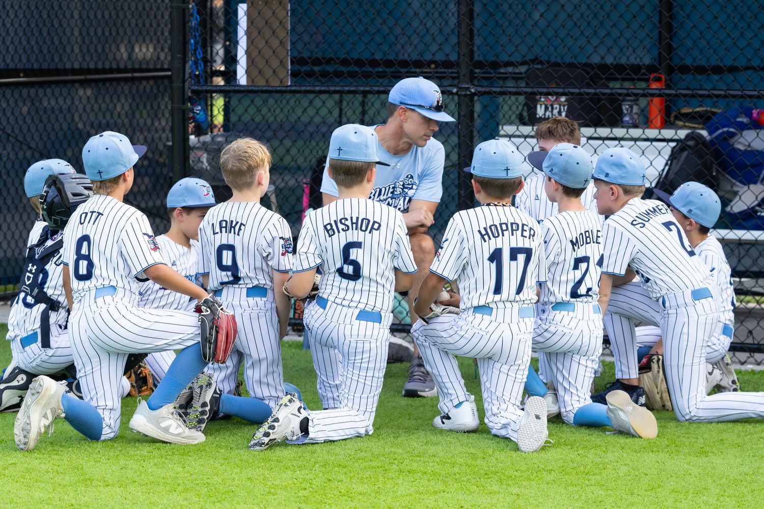 Youth baseball team huddles with coach in blue pinstripe uniforms during a Little League game.